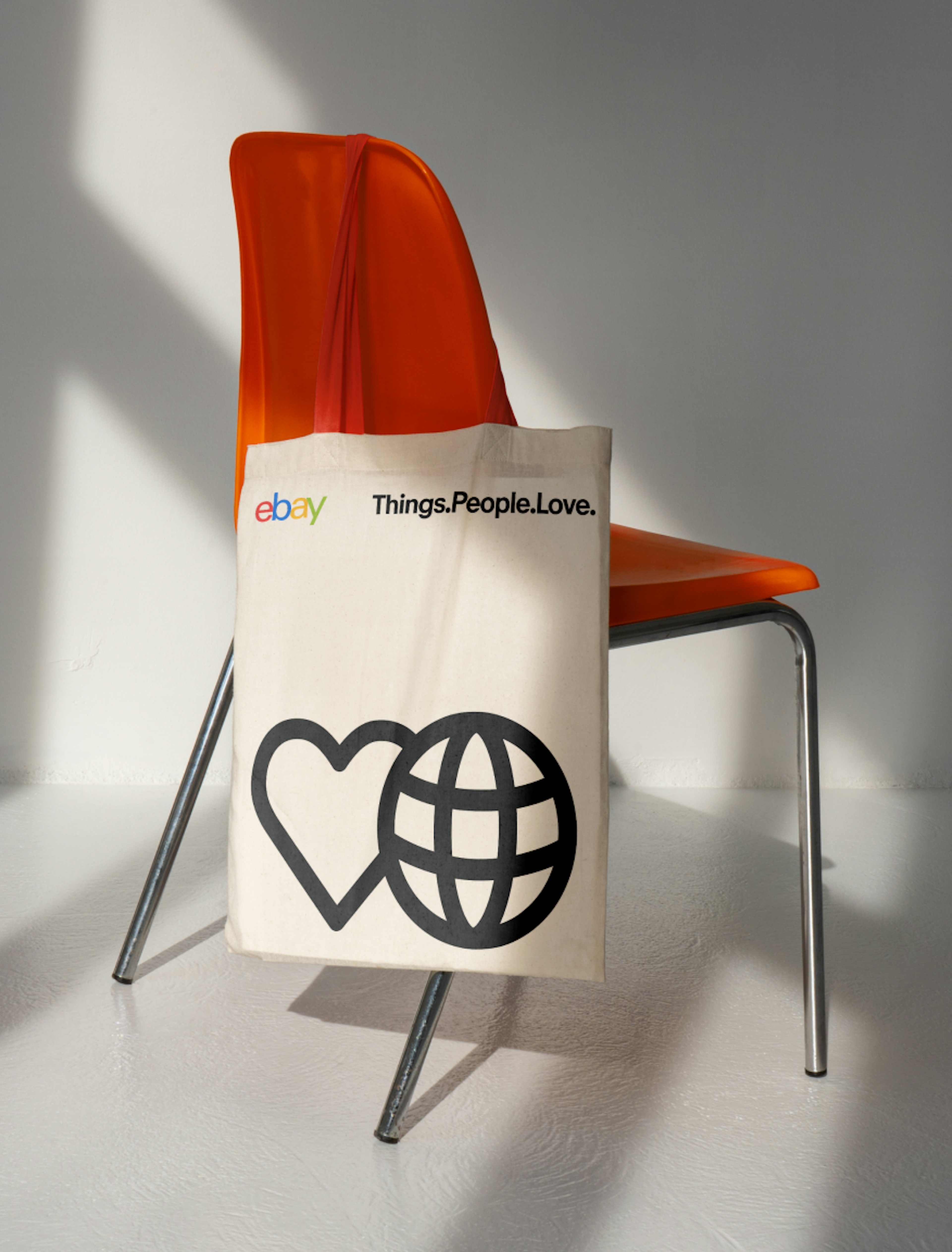 A white tote bag with the eBay logo and the text "Things People Love," featuring a heart and globe icon on the bottom. The tote bag is placed on an orange chair with a simple, modern backdrop.