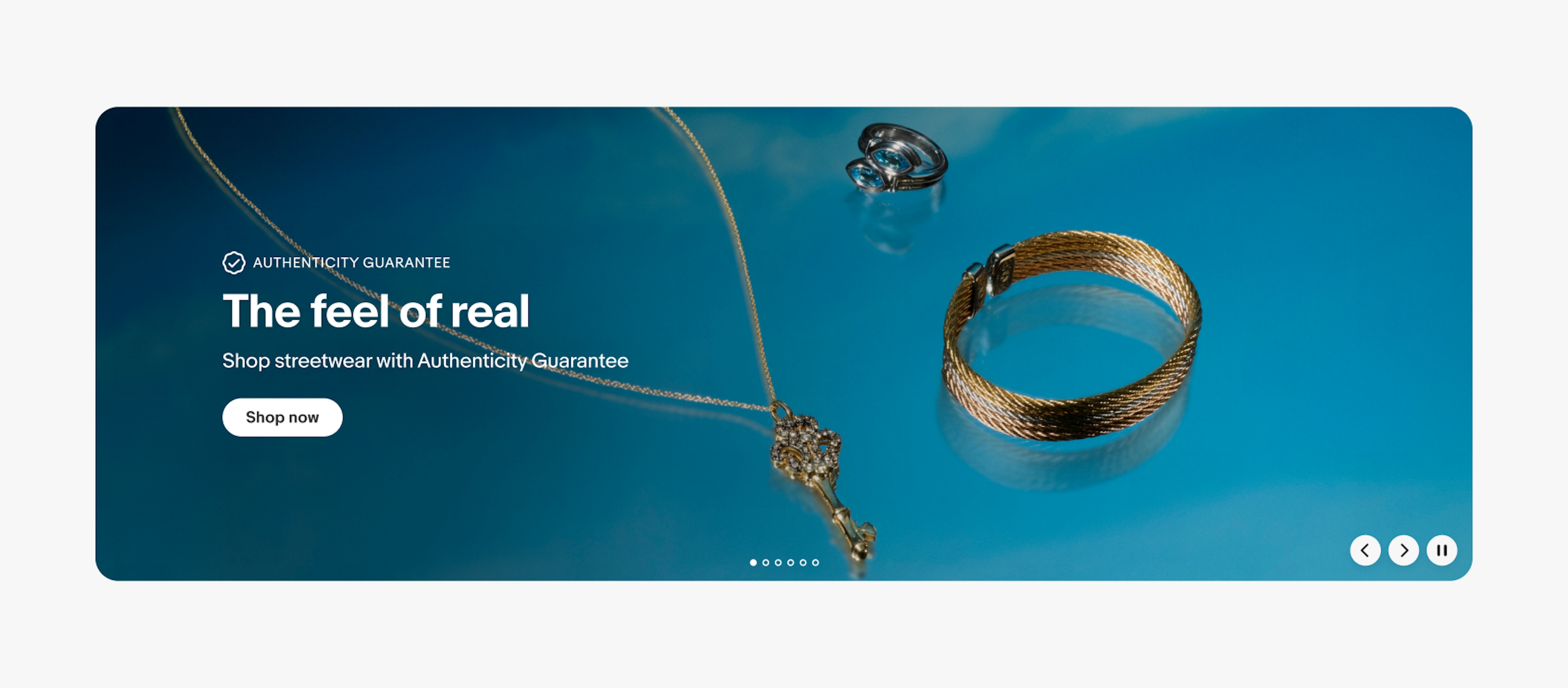 A large product banner with text on the left side containing an overline, headline, subline, and cta button. The image contains a vibrant blue background with 3 types of jewelry. The overline has the program lockup for Authenticity Guarantee.