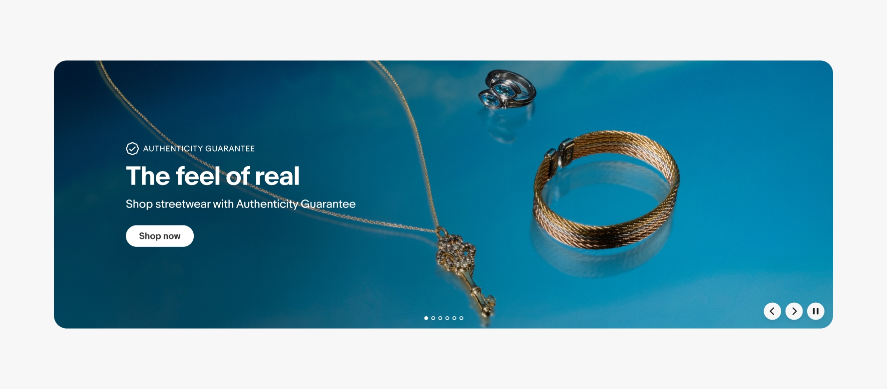 A large product banner with text on the left side containing an overline, headline, subline, and cta button. The image contains a vibrant blue background with 3 types of jewelry. The overline has the program lockup for Authenticity Guarantee.
