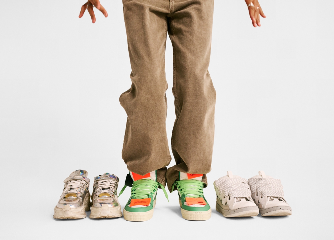 Three pairs of shoes neatly arranged next to each other on the ground. Someone wearing brown jeans is wearing the pair in the center.