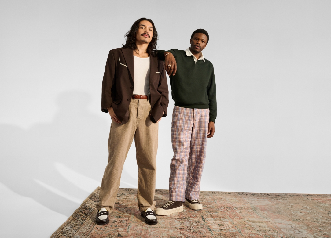 Two fashionable men standing next to each other on a vintage rug. The man on the right is resting his arm on the other man’s shoulder.