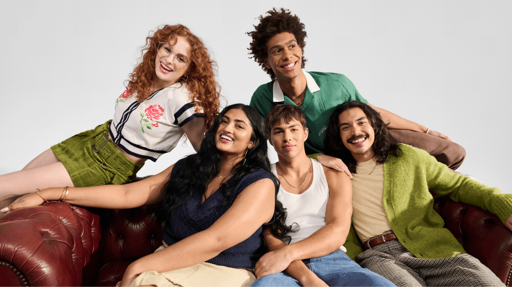 A diverse friend group of five on a brown leather couch wearing fashionable clothes.