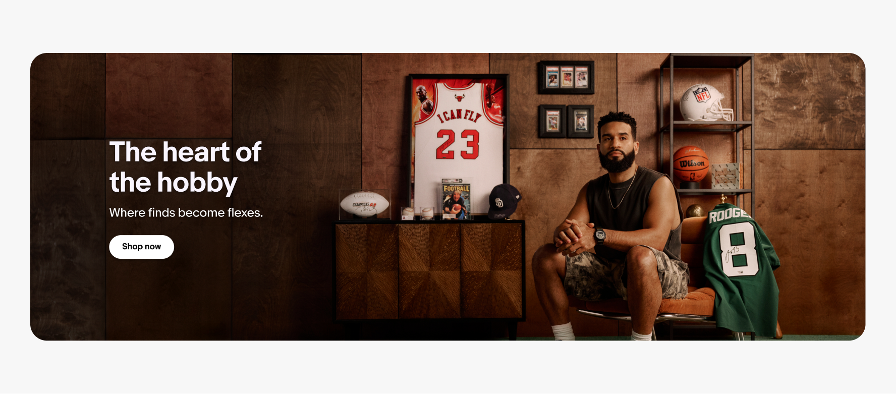 The image shows a man sitting in a chair next to a collection of sports memorabilia. The collection includes a framed basketball jersey with the number 23, a football, a basketball, and a football helmet, as well as framed trading cards and a comic. The text on the image reads, "The heart of the hobby. Where finds become flexes." There is also a "Shop now" button displayed on the left side of the image.