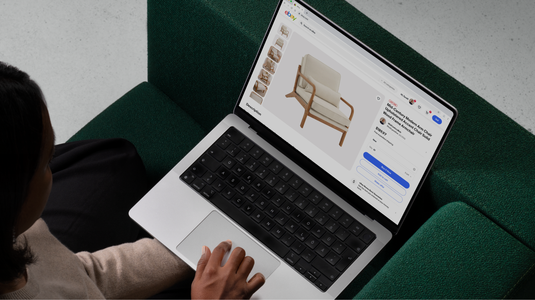 A woman holding a laptop sitting on a green couch. She is viewing a view item page on eBay containing a mid-century modern arm chair.