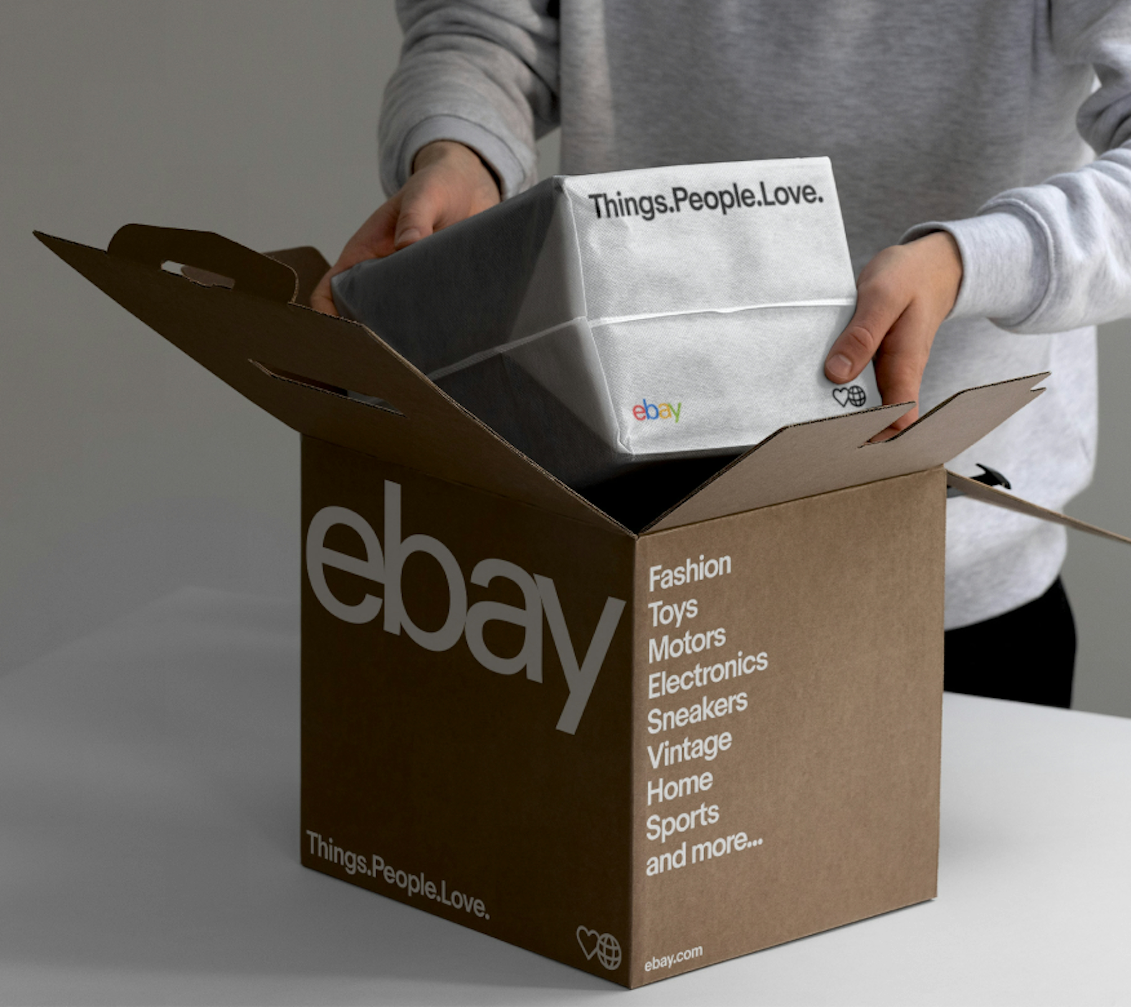 A person placing an item in a brown eBay box with white type on it. The Box has a large eBay logo, the tagline “Things.People.Love.” and the categories “fashion, toys, motors, electronics, sneakers, vintage, home, sports and more...” written out.