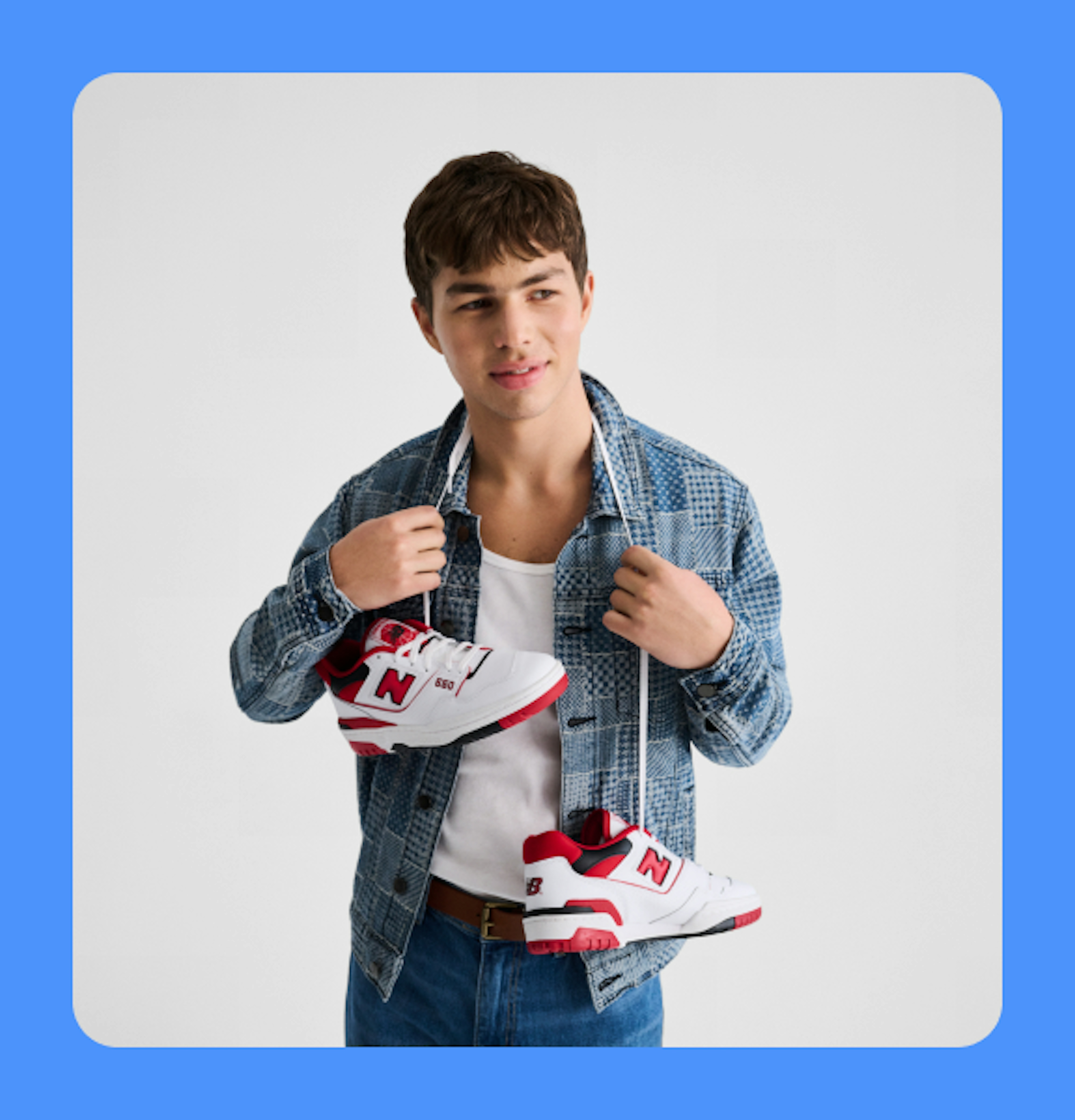 A lifestyle image of a person photographed against a white studio background wearing a jean jacket and jeans with red and white New Balance sneakers hanging around their neck by the laces, framed on a blue background.
