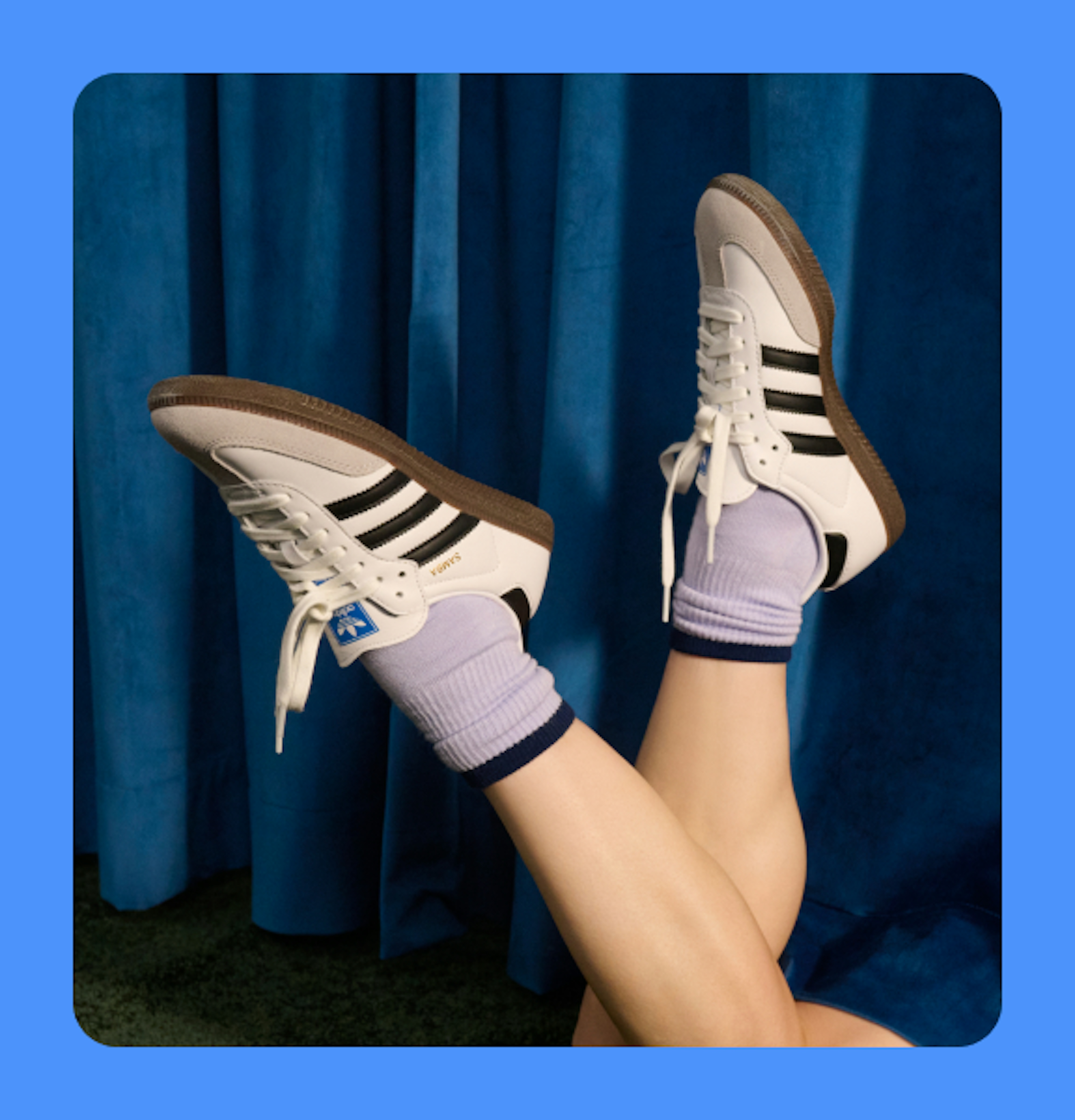 A studio closeup image of two legs, posed upwards wearing black-and-white Adidas sneakers and purple socks in front of a blue curtain, framed on a blue background.