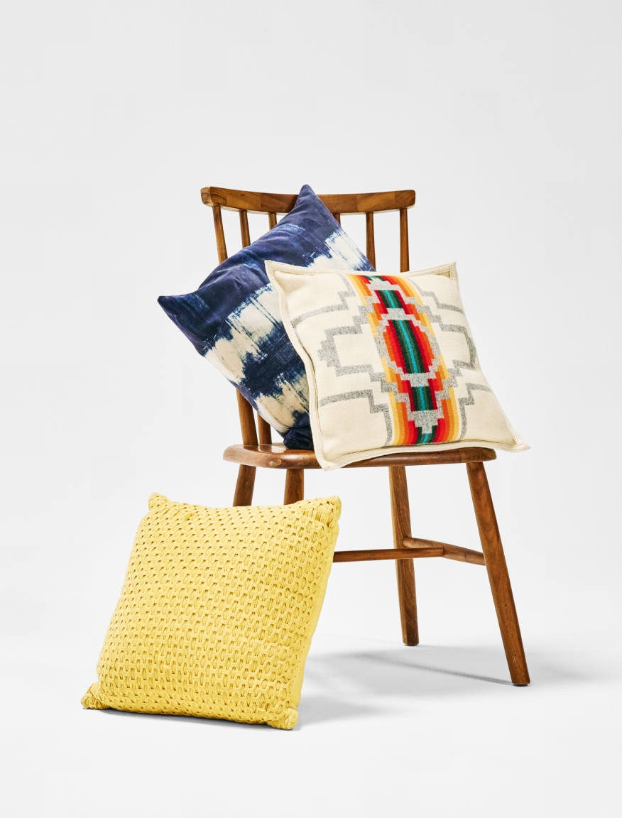 A wooden chair with two colorful decorative cushions on top of the chair, and one yellow one leaning on the chair leg, against a plain white background.