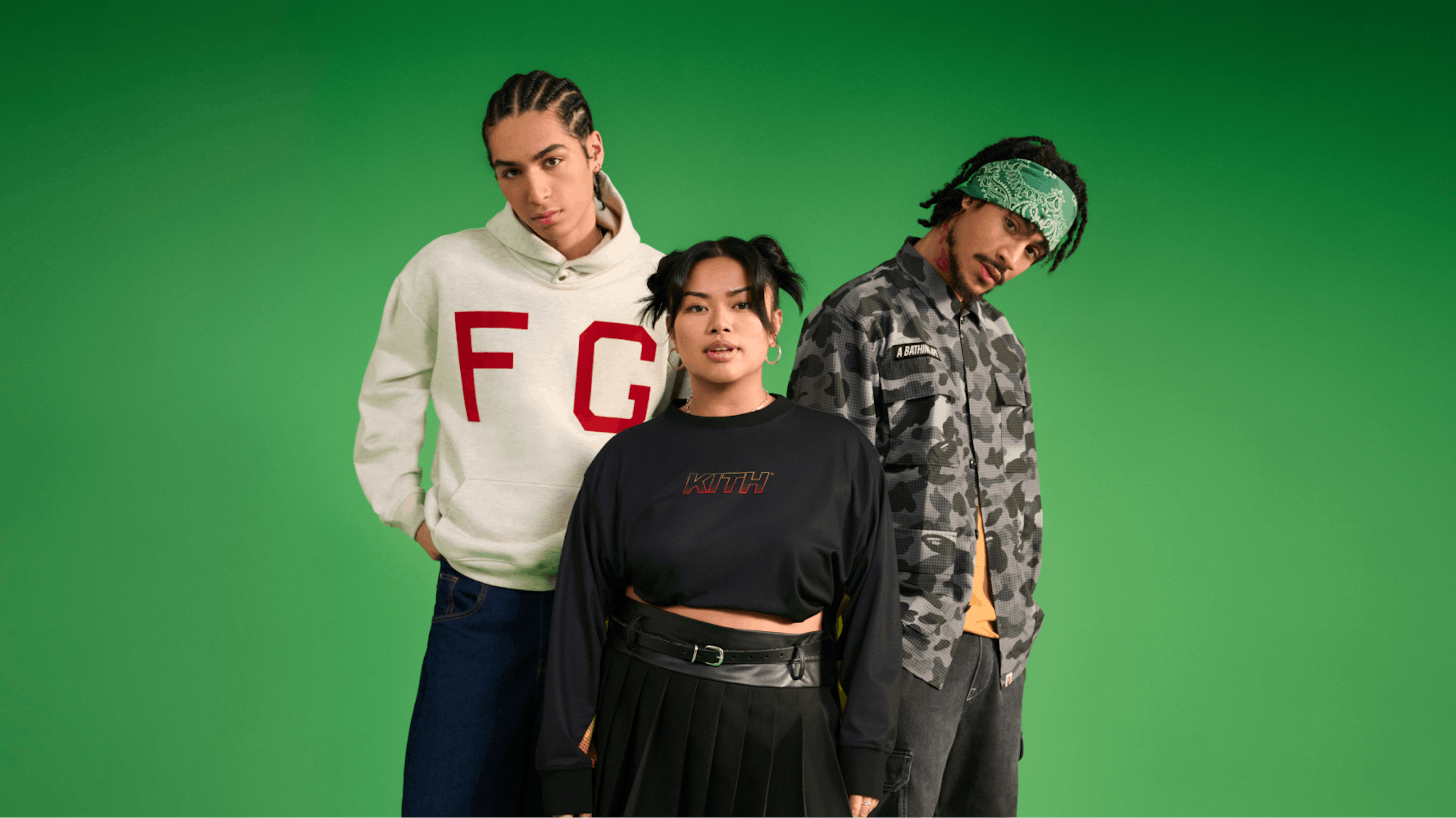 Three stylish young adults pose confidently against a green background. One wears a white hoodie with "F G" and dark jeans, another a black cropped "KITH" sweatshirt with a pleated skirt, and the third a camouflage jacket from A Bathing Ape brand, green bandana, and dark pants. Their styles blend casual and streetwear fashion.