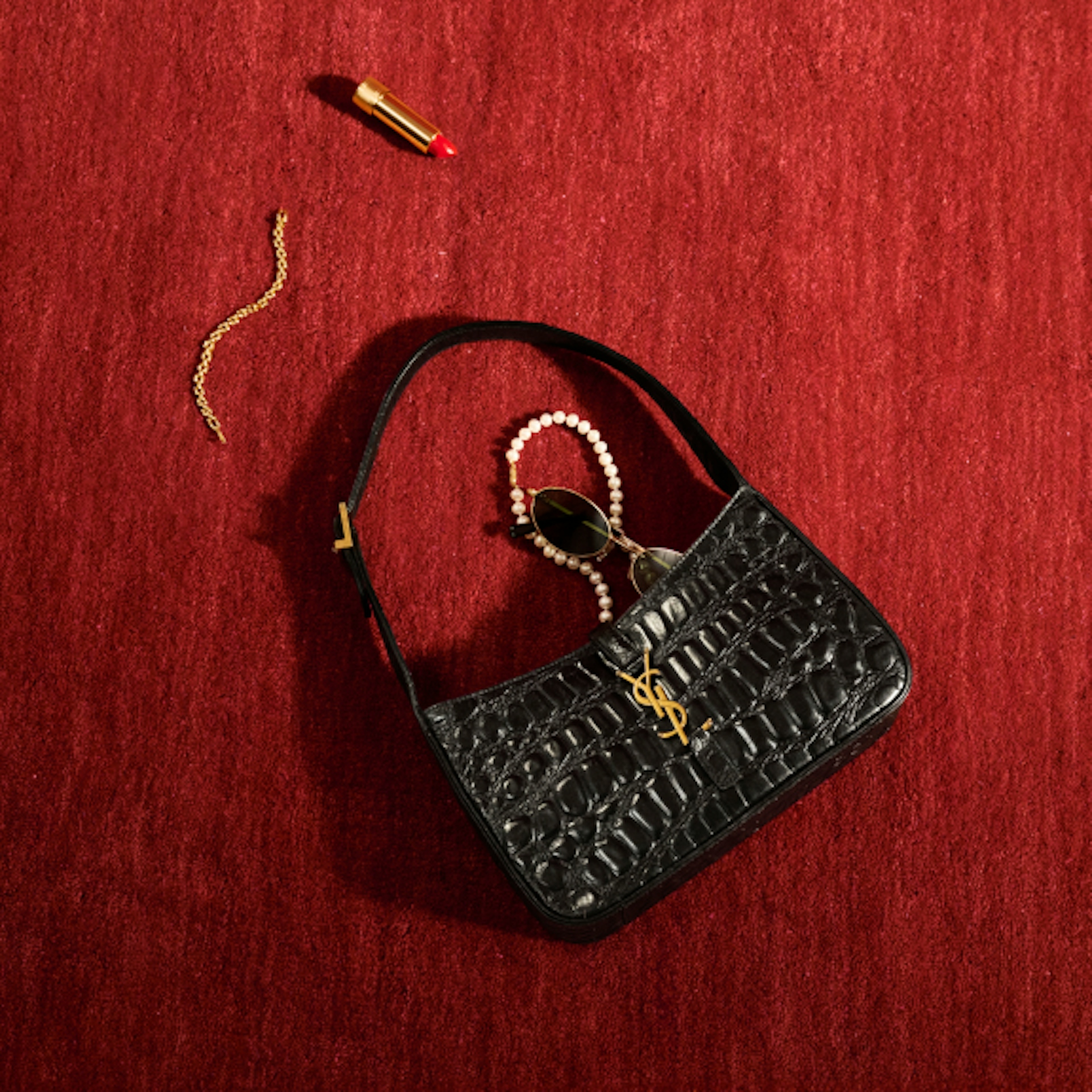 A black croc-embossed YSL handbag is placed on a red textured surface. Nearby, there are a red lipstick, a gold chain, and a pair of sunglasses with a pearl chain. The arrangement showcases a chic and luxurious aesthetic.