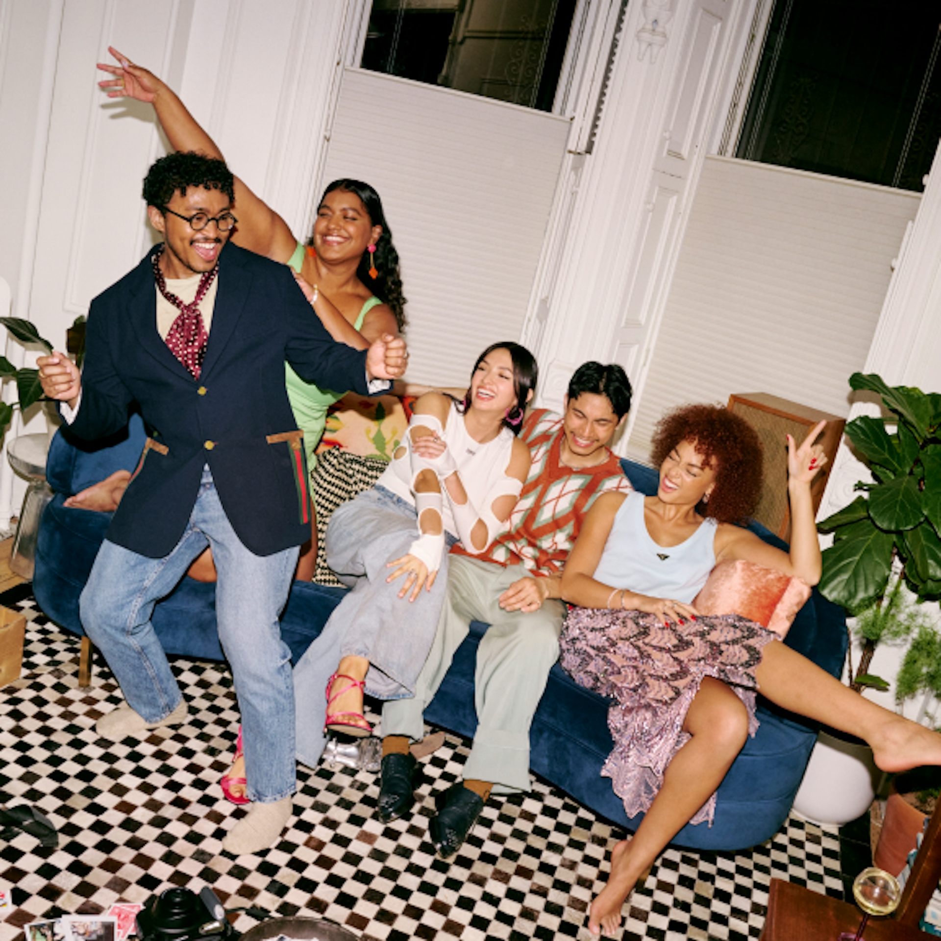 The image depicts five friends enjoying themselves indoors, sitting on a blue couch in a stylish room with white walls and large windows. The floor features a black and white checkered pattern. The group appears joyful, with some dancing and others laughing, dressed in colorful and trendy outfits. The room is decorated with plants and has a casual, lived-in feel.
