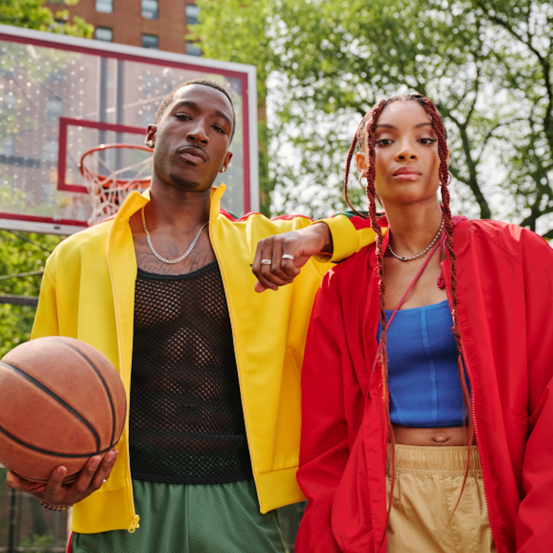 The image shows two individuals standing confidently on a basketball court outdoors. The person on the left is holding a basketball and wearing a yellow jacket over a black mesh shirt and green shorts. The person on the right is wearing a red jacket over a blue top and beige pants. Both have a stylish and casual appearance, with a backdrop of trees and a basketball hoop visible behind them. They exude a cool, urban vibe with their relaxed yet assertive postures.