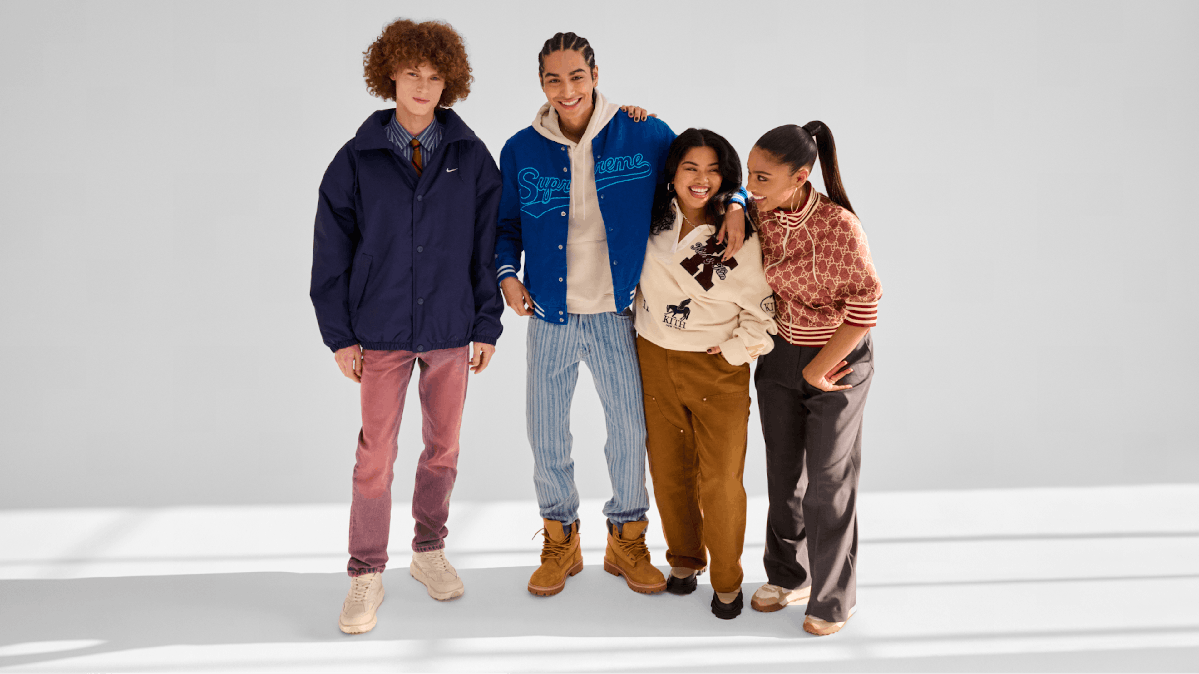 The image shows four young individuals dressed in a mix of athleisure and streetwear styles. A description of the looks: On the left, we see a dark blue jacket over a striped collared shirt, pinkish-red faded trousers, and beige sneakers. On the second Left: Blue varsity jacket with "Supreme," light-colored hoodie, light blue striped jeans, and tan Timberland-style boots. On the second right, we see a cream sweatshirt with "KITH" and graphic print, mustard brown cargo pants, and black shoes. And lastly, on the far right we see a Burgundy patterned zip-up top, dark gray flared trousers, and beige sneakers.