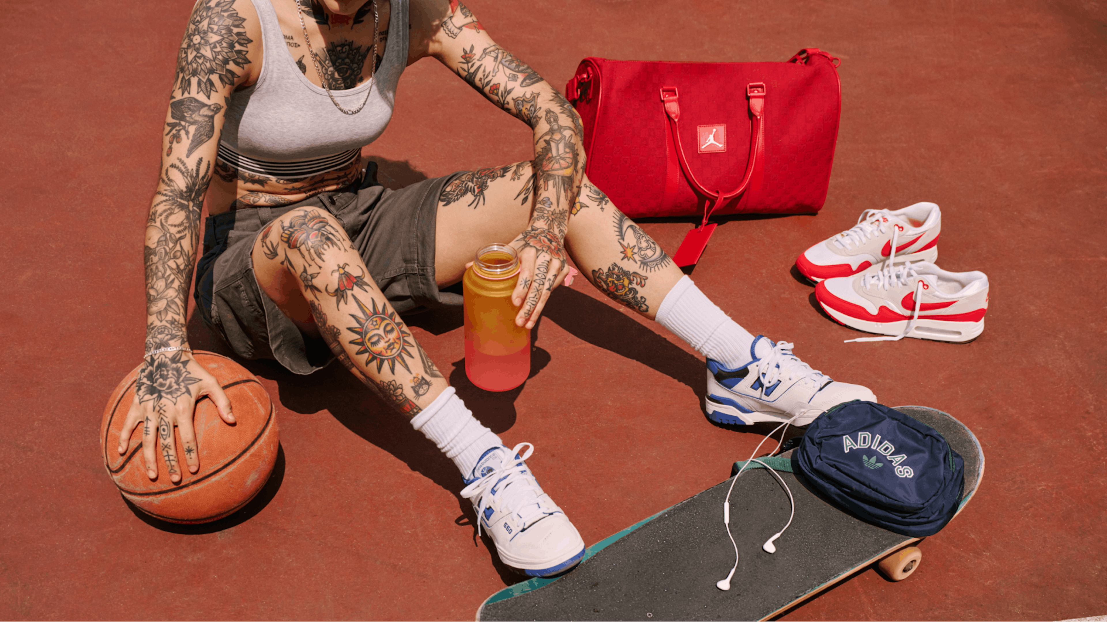 The image shows a person with numerous tattoos on their arms and legs sitting on a red surface, likely an outdoor court. They are wearing a gray sports bra, gray shorts, white socks, and blue and white sneakers. A basketball rests between their legs, and they hold a gradient water bottle. Beside them are a red sports bag, a pair of red and white sneakers, a blue Adidas pouch, and a skateboard with white earphones. The scene exudes a sporty and casual vibe.