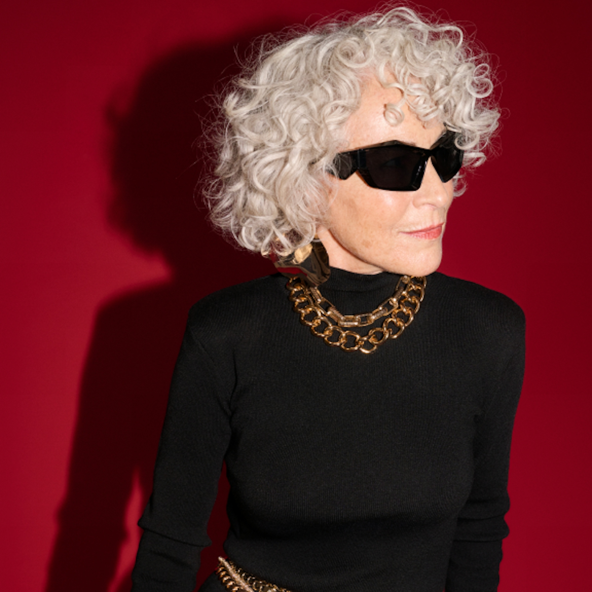The image shows an older woman with curly white hair, wearing large black sunglasses and a fitted black turtleneck. She accessorizes with bold chunky chain necklaces and oversized earrings. The background is a solid deep red, highlighting her stylish, sophisticated appearance.