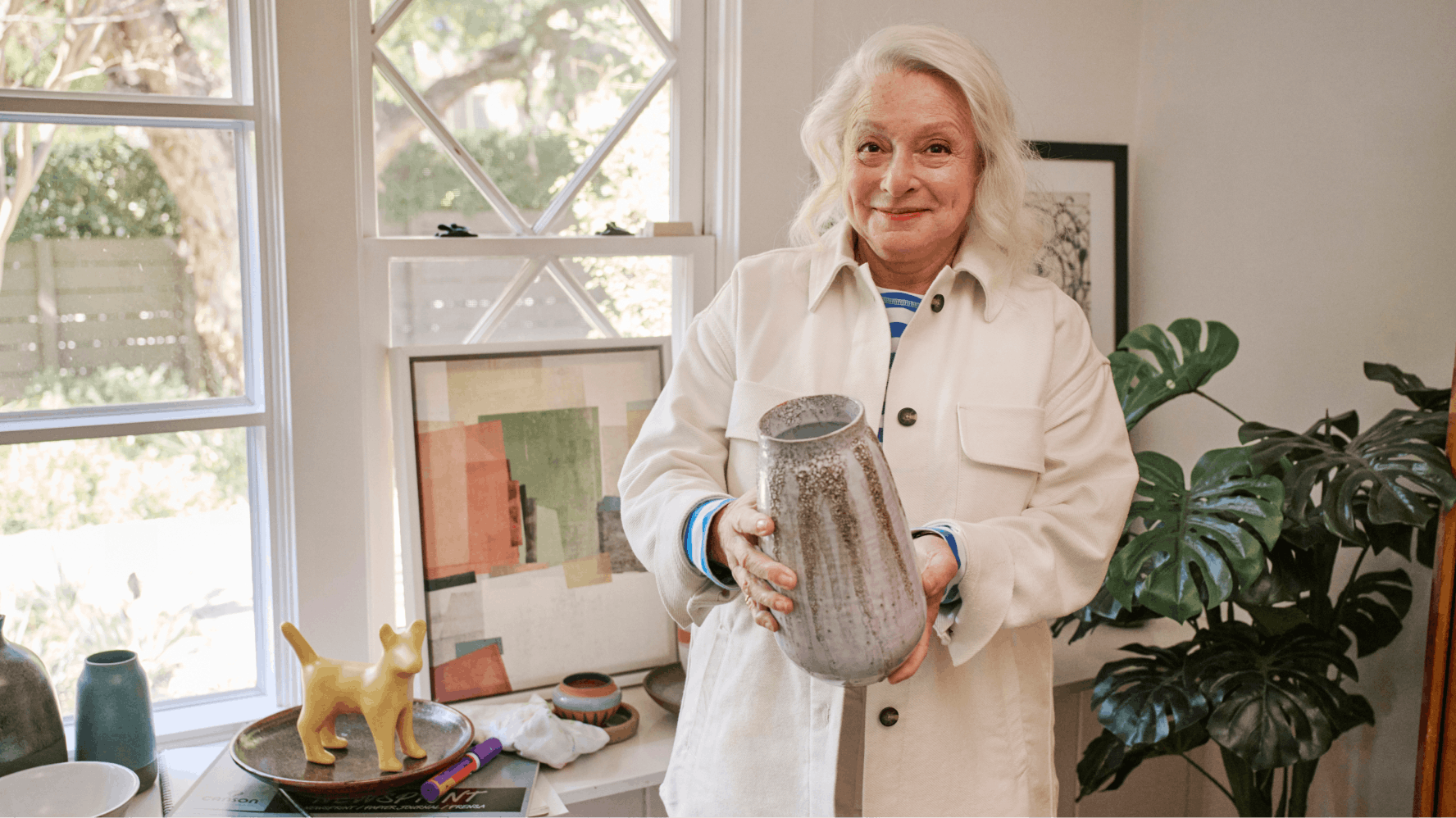 The image shows an older woman standing indoors, holding a large, textured vase. She is wearing a light-colored jacket over a blue and white striped shirt. The background features a bright room with large windows, artwork, a potted plant, and a table with various decorative items. The scene conveys a cozy, artistic atmosphere.