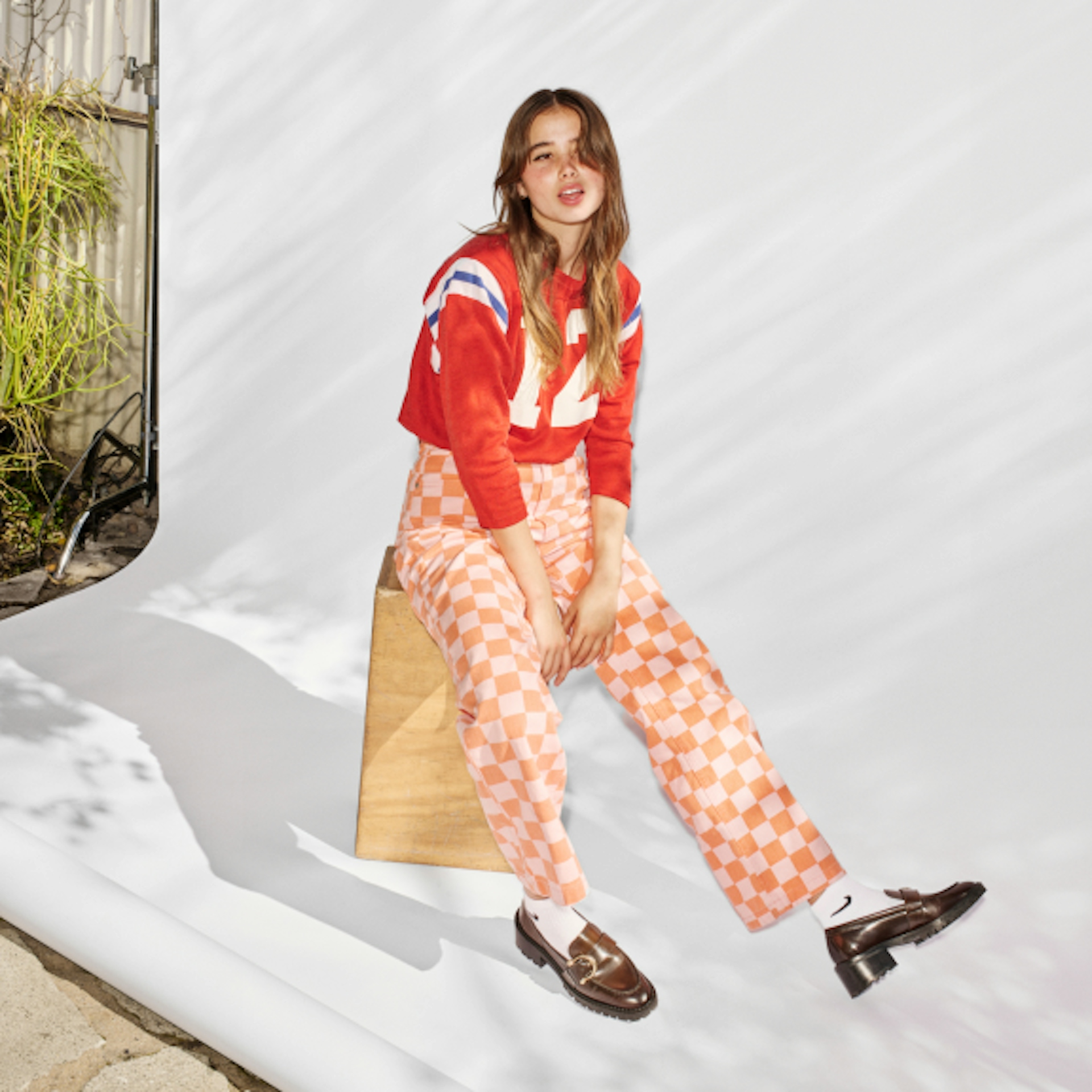 The image shows a young woman seated on a wooden block, dressed in a red cropped sweatshirt with white and blue stripes and the number "72." She pairs it with high-waisted, pink and white checkered pants. Her outfit is completed with white socks and brown loafers. The background is a bright, white backdrop with some natural outdoor elements visible on the left side.