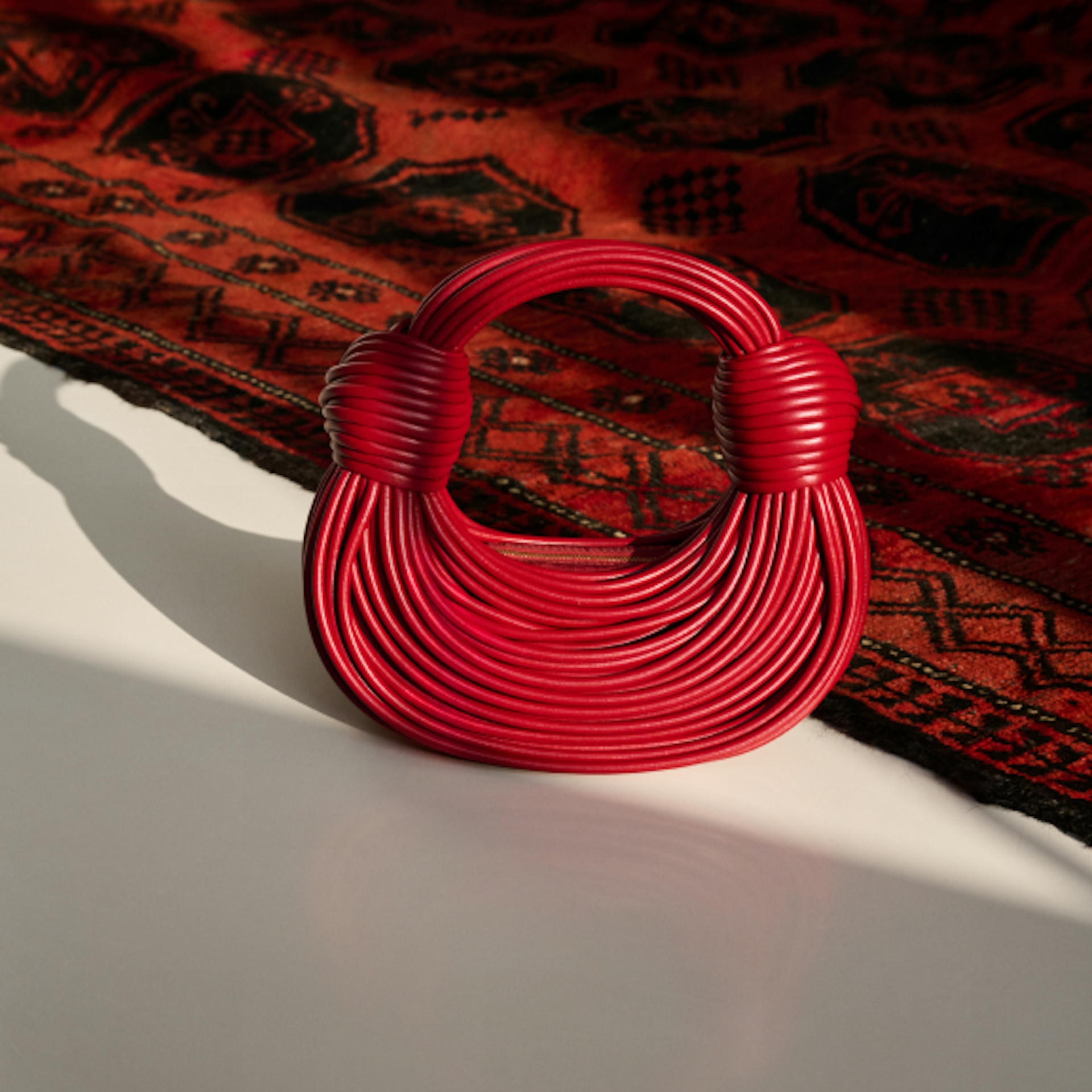 The image shows a stylish, small red handbag with a unique, coiled design. It is placed on a surface with a richly patterned red and black rug in the background, highlighting the bag's intricate texture and bold color.