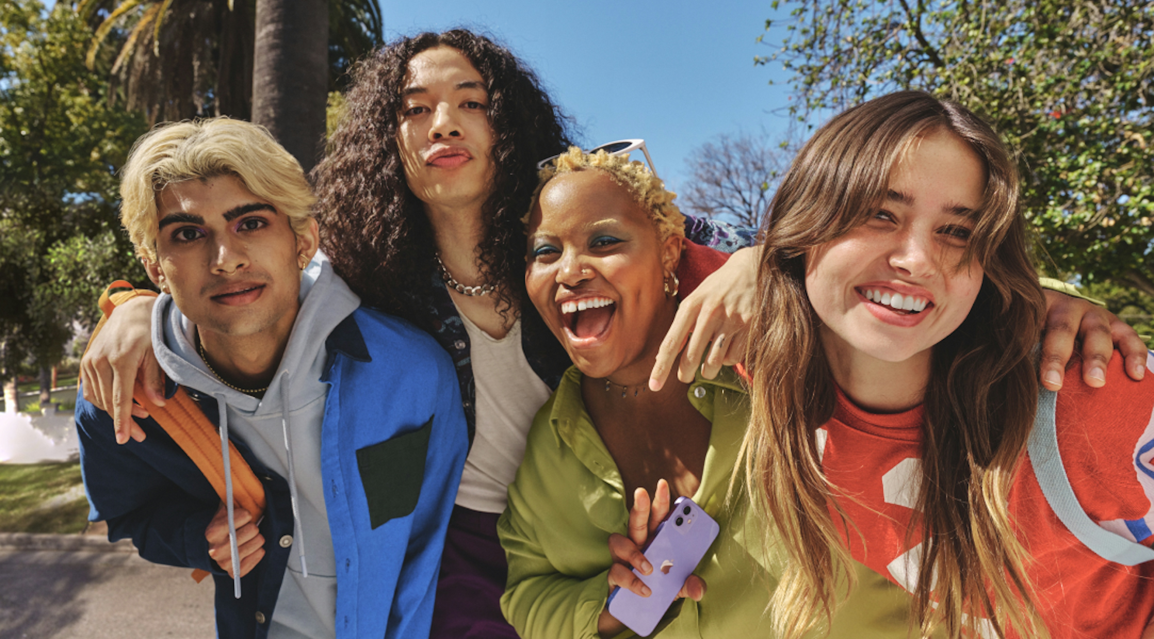The image shows four young adults closely huddled together, smiling and happy. They are dressed in casual, colorful clothing and are outdoors on a sunny day. One person is holding a smartphone, and they all seem to be enjoying each other's company.