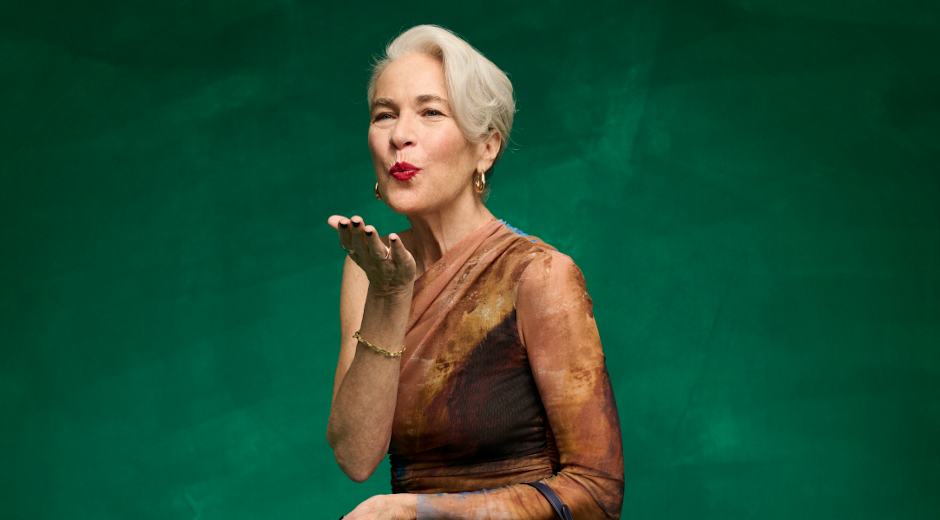 The image shows an older lady dressed in a brown casual dress situated in a green painted studio background. She is blowing a kiss to the camera.