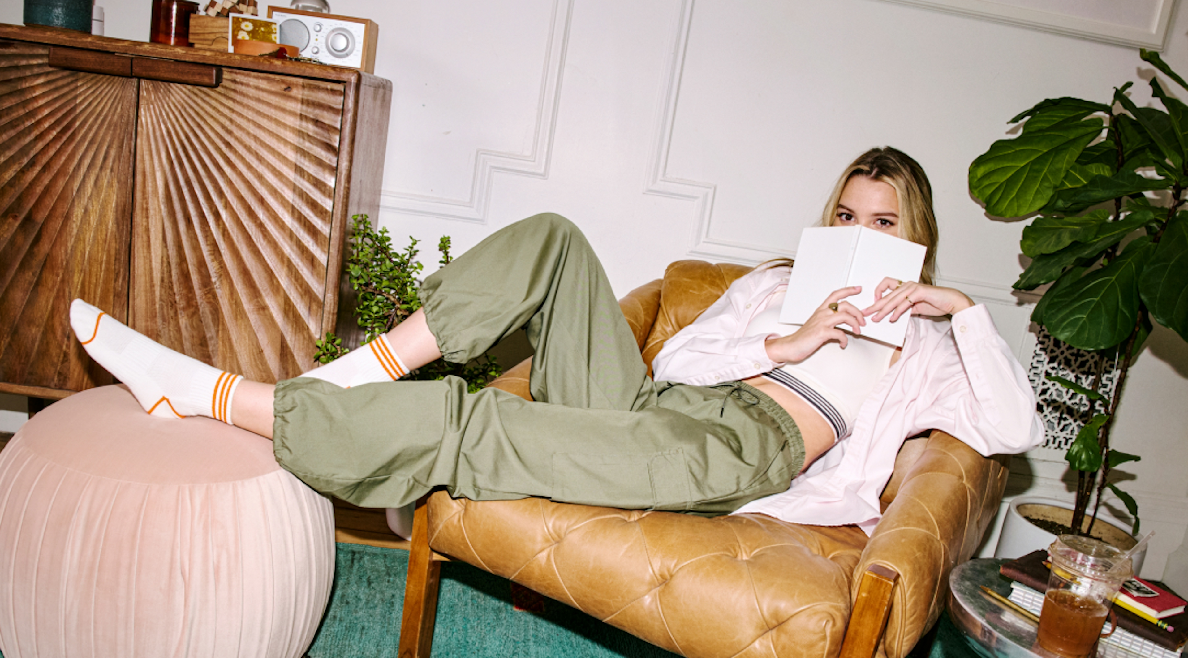 The image shows a young lady sitting very relaxed on a leather armchair in her living room at home. She is playfully hiding her face behind the book she is reading.