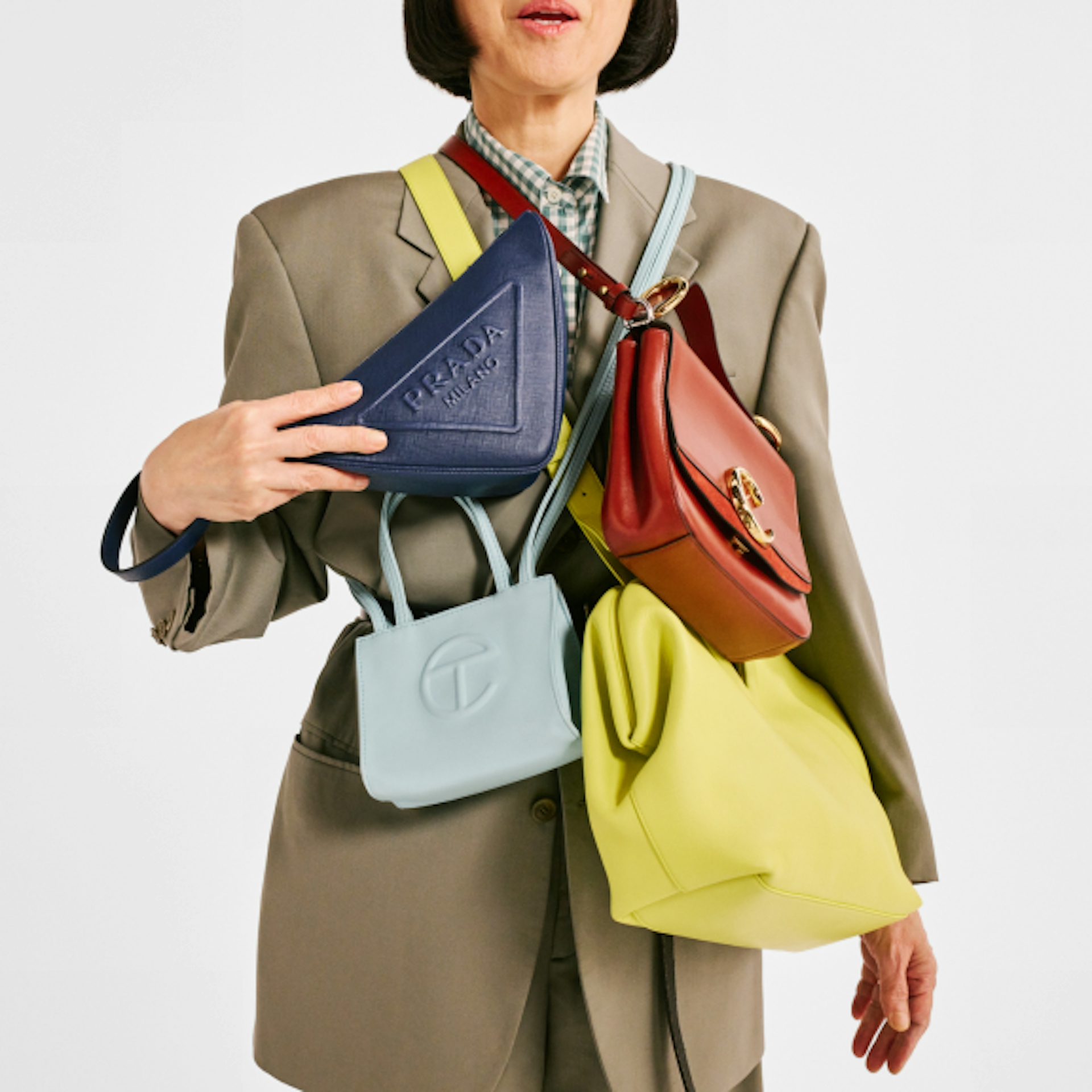 A person dressed in a beige suit is holding multiple handbags of various colors and styles, including a blue Prada bag, a light blue Telfar bag, a red handbag, and a yellow bag. The image highlights the variety and fashion-forward nature of the bags.