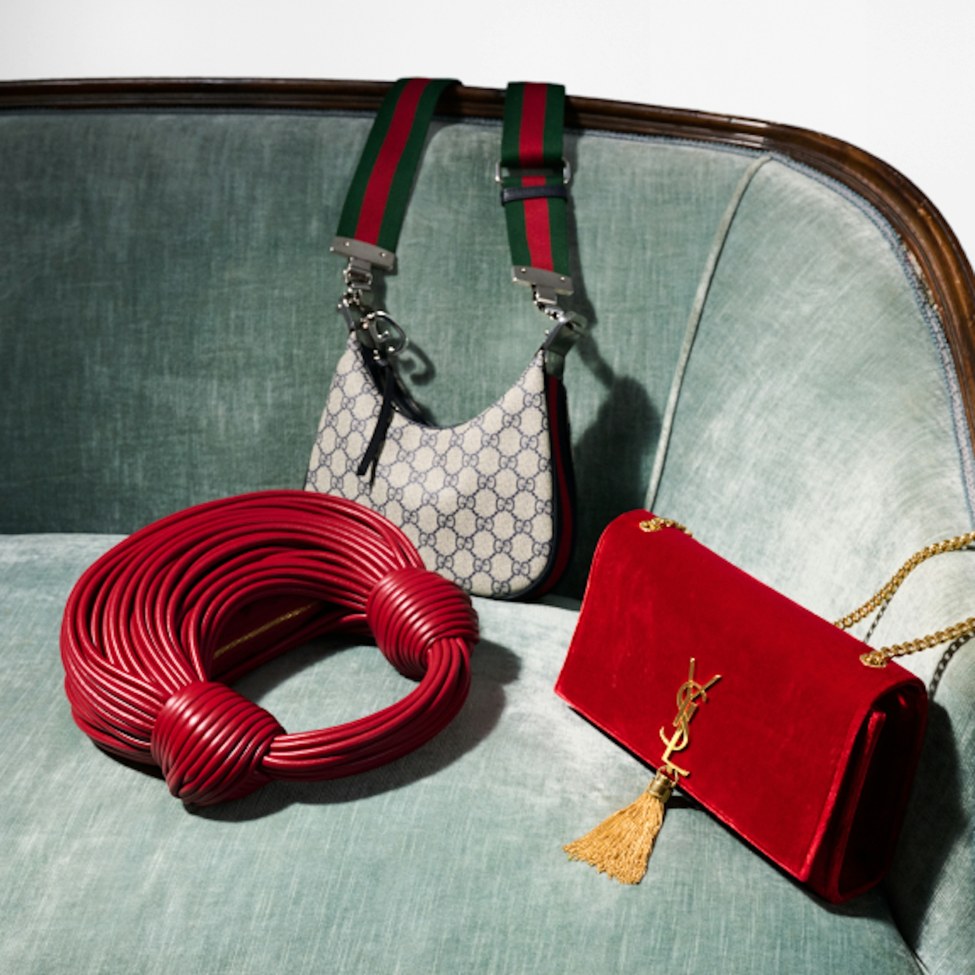 The image displays three stylish handbags arranged on a green velvet sofa. One is a white Gucci bag with green and red striped straps, the second is a unique red coiled design, and the third is a red YSL bag with a gold tassel and chain strap.