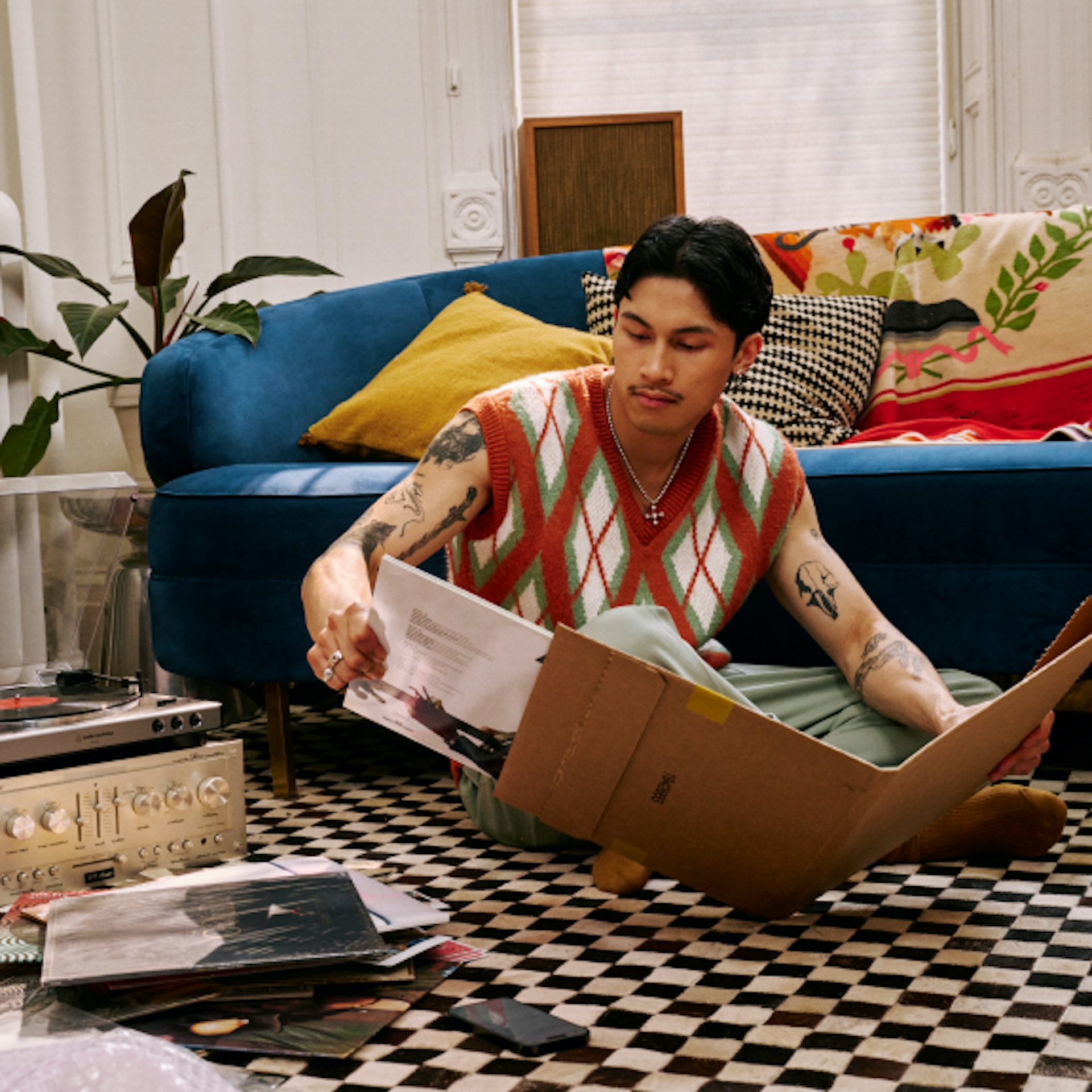 A person is sitting on the floor of a cozy living room, looking through vinyl records. They are wearing a colorful argyle vest and green pants, with a blue couch, patterned pillows, and plants in the background. The scene has a vintage and relaxed vibe.