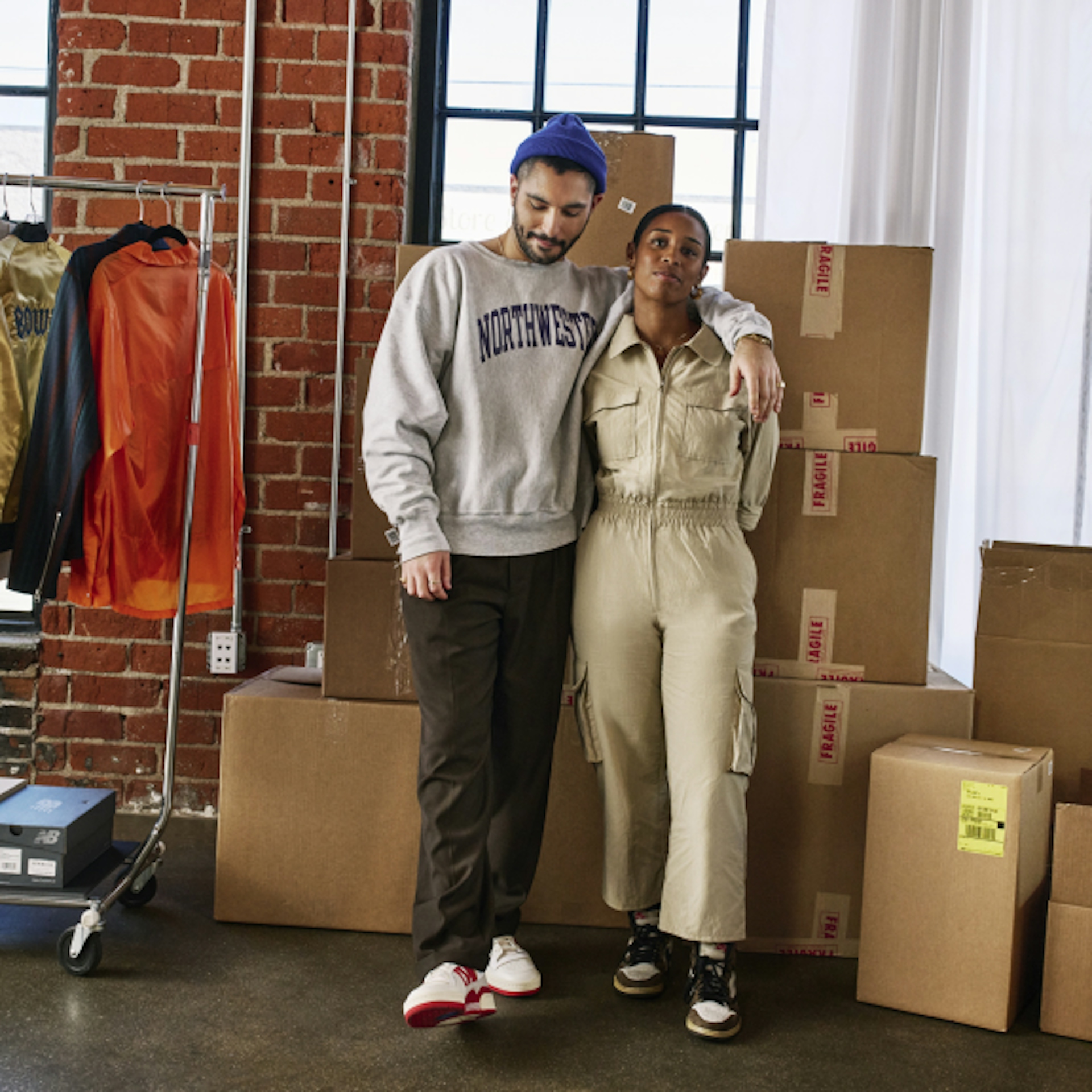 Two people stand close together in a room filled with cardboard boxes. One is wearing a grey "Northwestern" sweatshirt and a blue beanie, while the other is dressed in a beige jumpsuit. The background features a brick wall and a clothing rack with various garments, suggesting a casual and industrious setting.