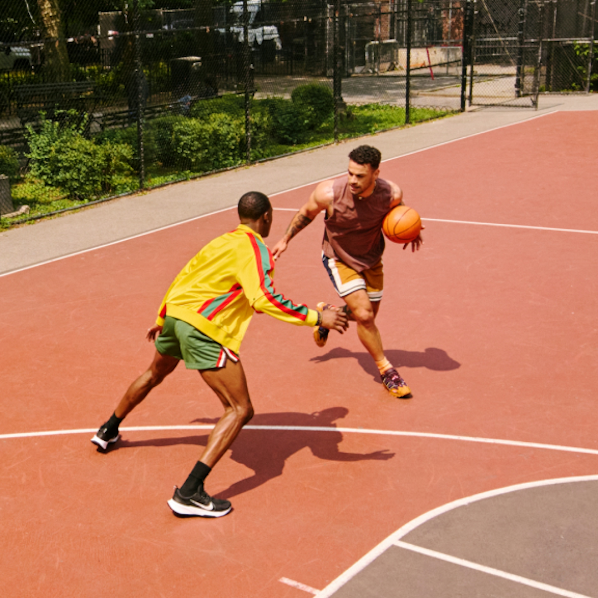 Two people are playing basketball on an outdoor court. One, wearing a yellow jacket and green shorts, is defending against the other, who is dribbling the ball while wearing a brown sleeveless shirt and multicolored shorts. The court is surrounded by a fence and greenery.