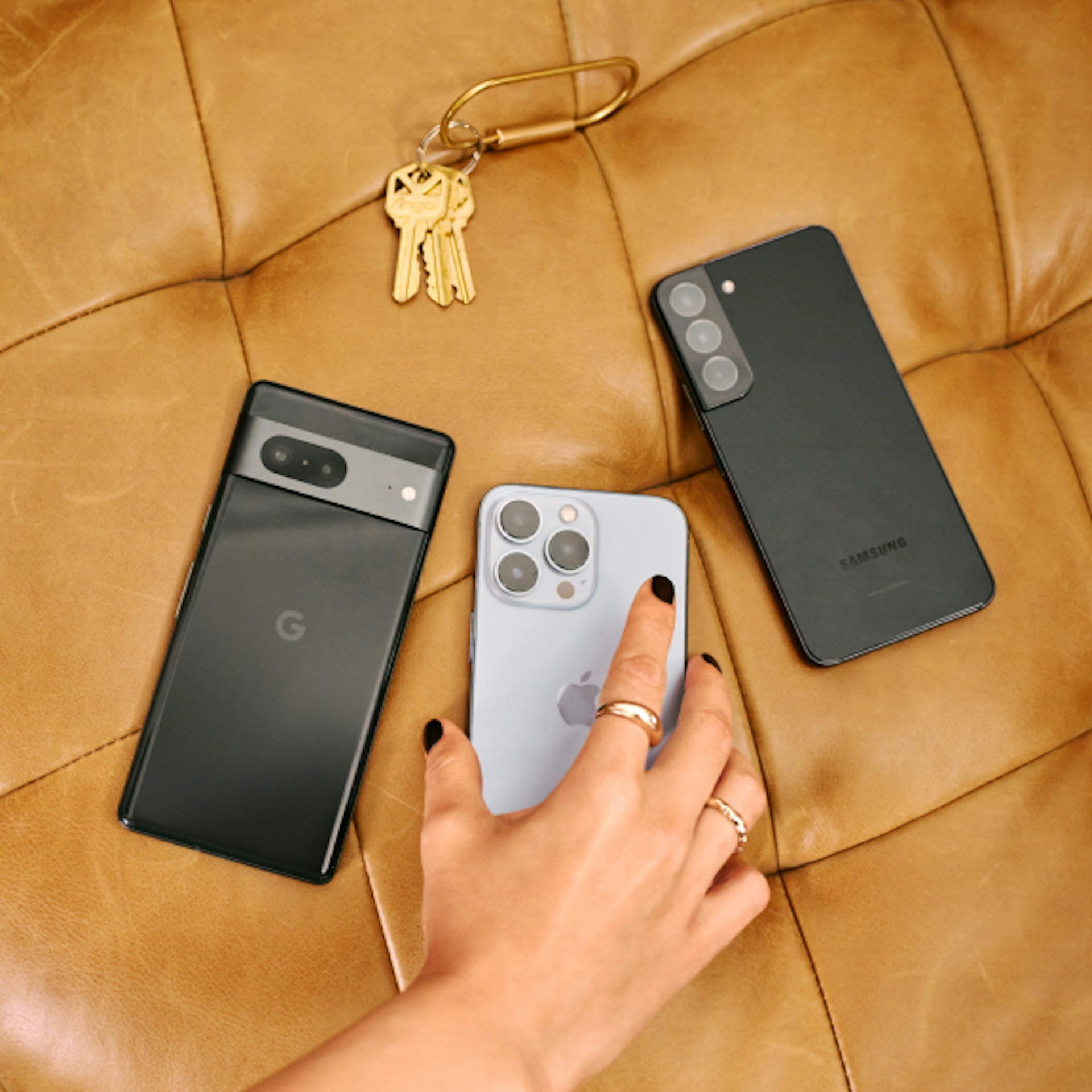 Three smartphones (Google, Apple, and Samsung) are placed on a tan leather surface alongside a set of keys. A hand with black nail polish and gold rings is reaching for the iPhone.