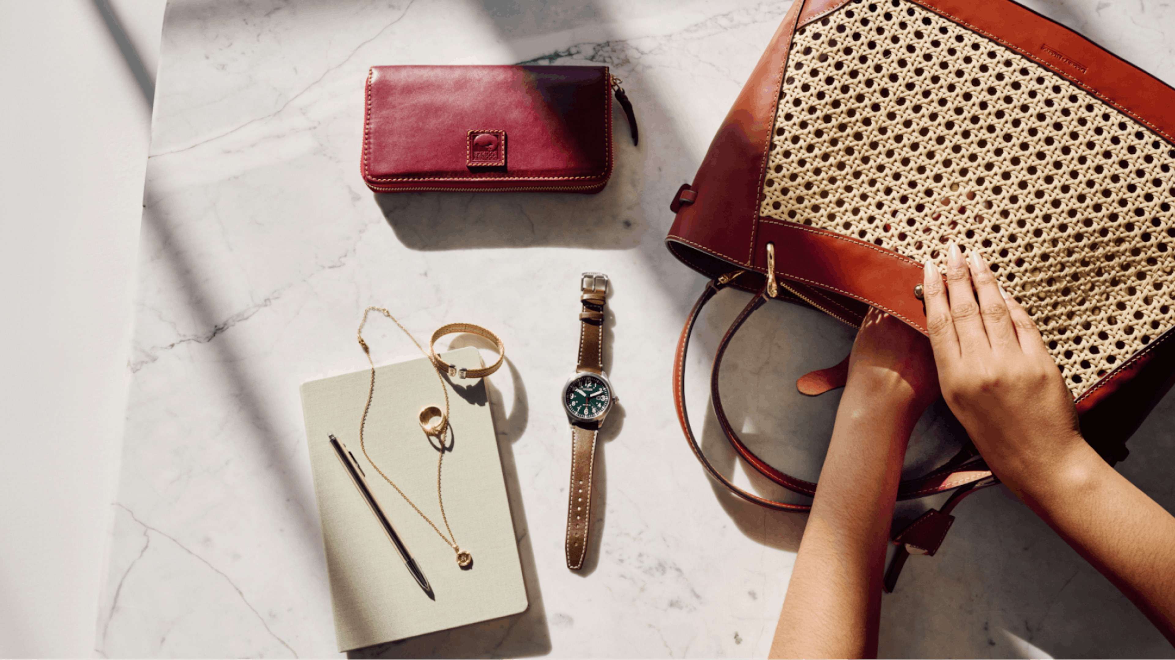 A neatly arranged collection of items is displayed on a marble surface. It includes a red leather wallet, a green-faced watch with a brown strap, a gold bracelet, a gold ring, a necklace, a notebook with a pen, and a woven handbag with leather trim. A pair of hands is seen reaching into the handbag.