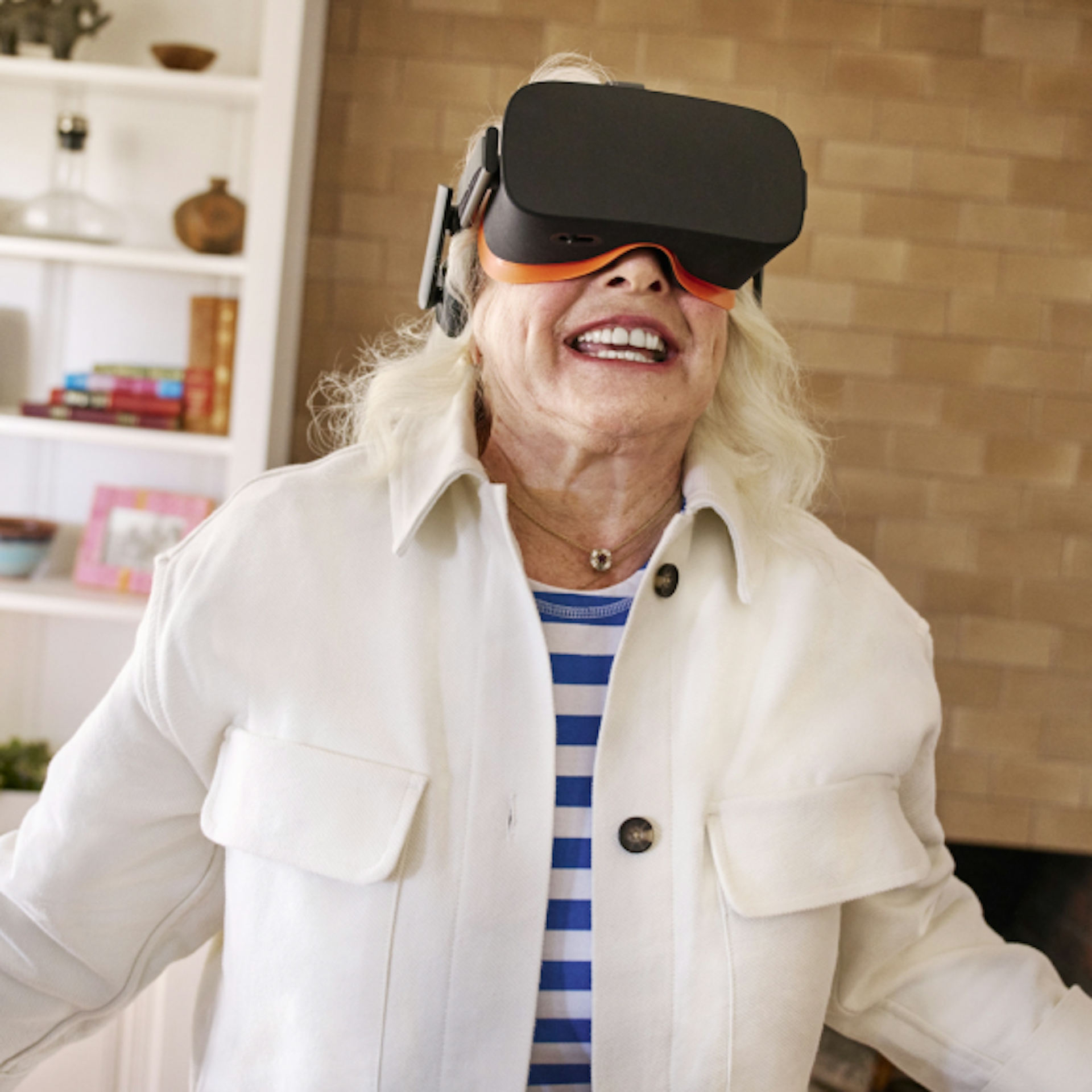 An older woman is enjoying a virtual reality experience, wearing a VR headset. She is dressed in a white jacket over a blue and white striped shirt, smiling with excitement. The background shows a cozy room with bookshelves and a brick wall.