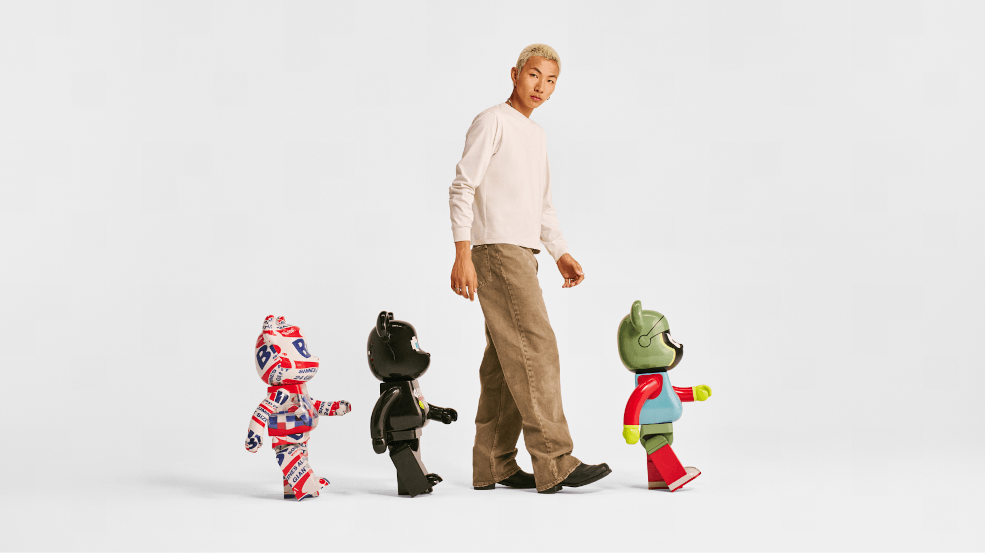 A person with bleached blonde hair, wearing a light long-sleeve shirt and beige pants, is walking alongside three colorful bearbricks. The bearbricks are designed with distinct patterns and colors, creating a playful and whimsical scene against a plain white background.