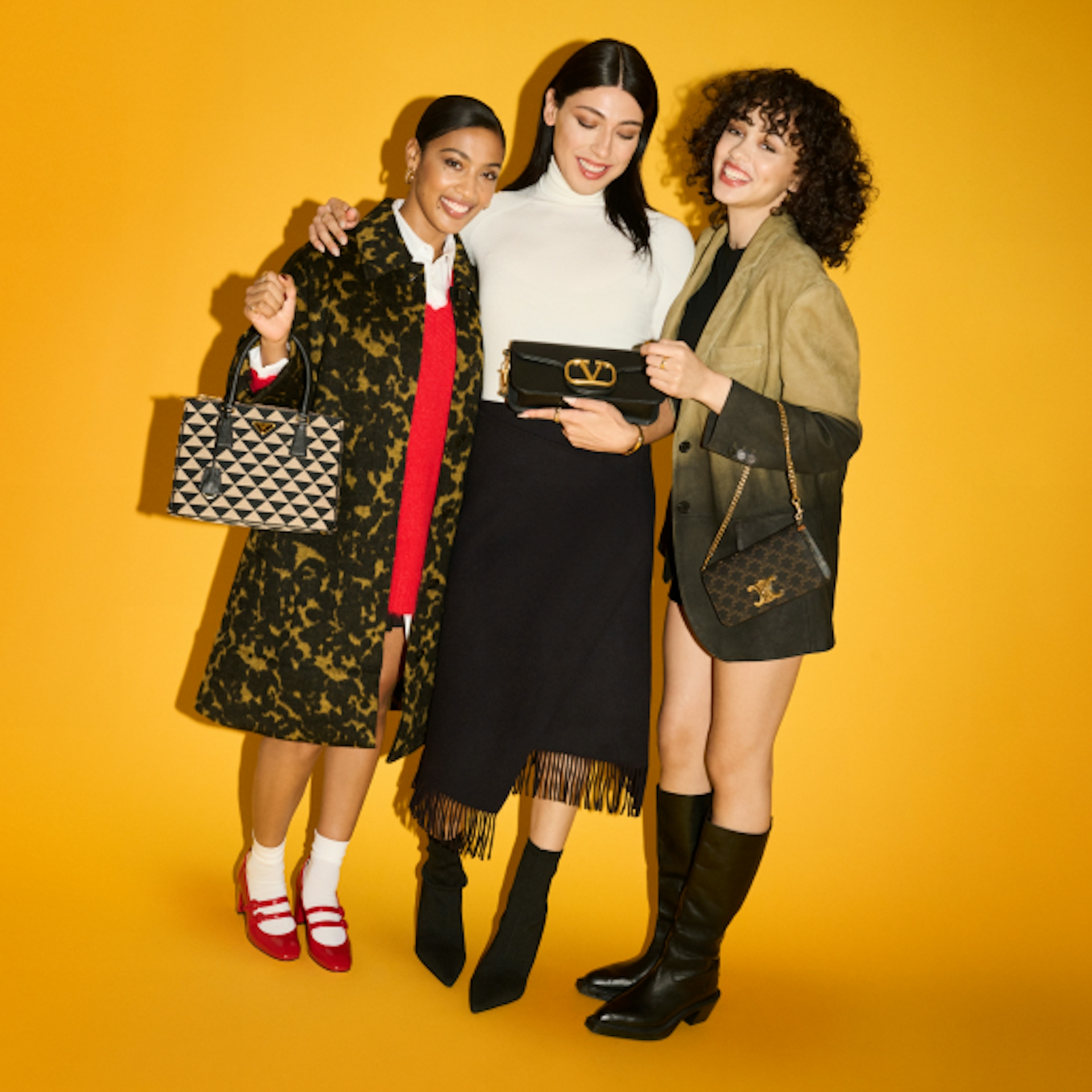 Three women are standing together against a yellow background, smiling and holding designer handbags. They are dressed stylishly: one in a patterned coat with a red dress, another in a white turtleneck and black skirt, and the third in a green jacket and black boots. They exude confidence and elegance.