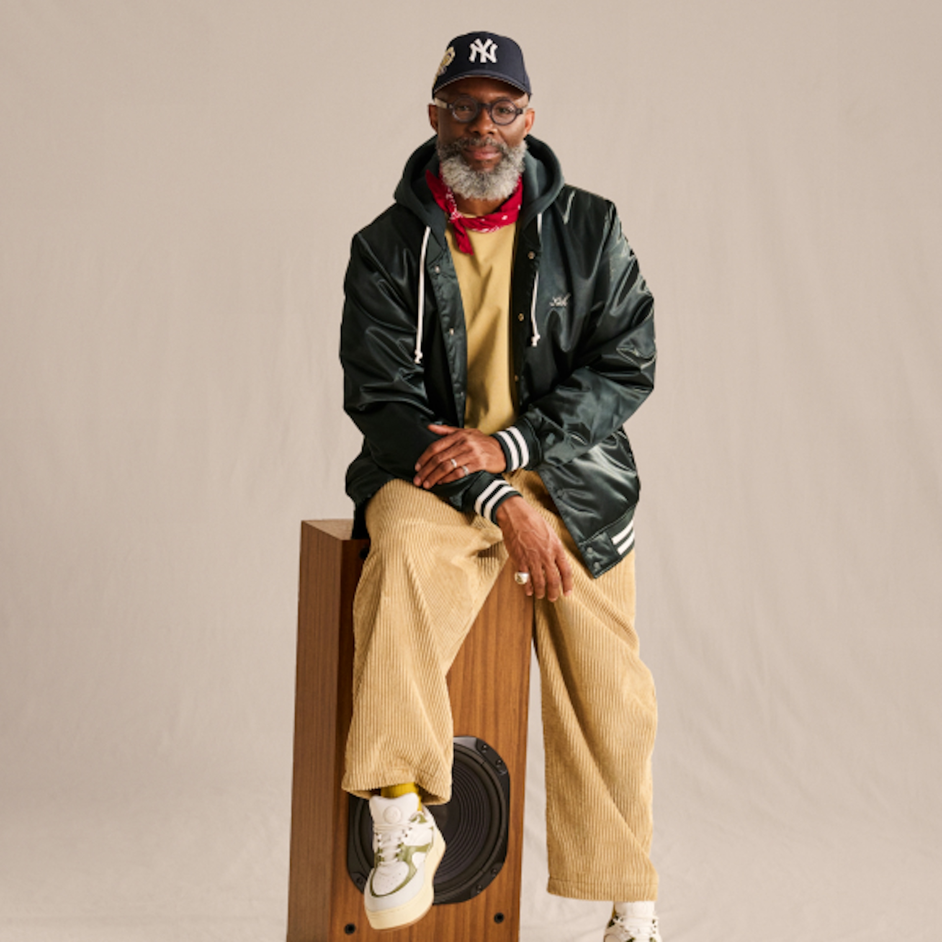 An older man with a beard is sitting on a wooden speaker. He is wearing a black baseball cap, glasses, a red bandana, a black jacket, a beige sweatshirt, and beige corduroy pants. He exudes a casual and stylish vibe.