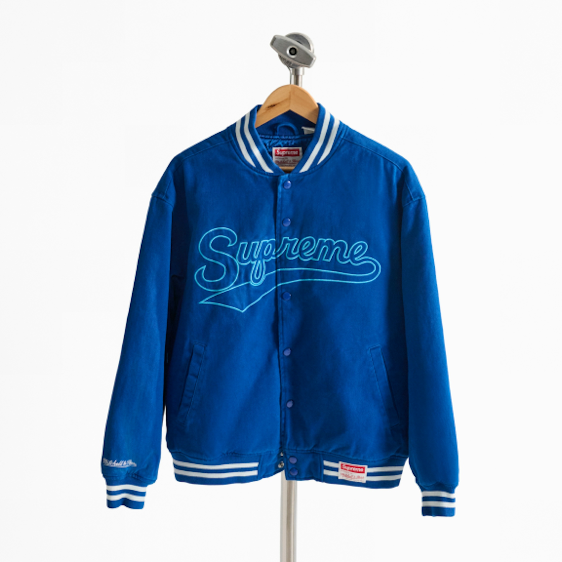 A blue Supreme varsity jacket is displayed on a hanger. The jacket features the word "Supreme" written in a cursive style on the front, with white striped accents on the collar, cuffs, and hem.