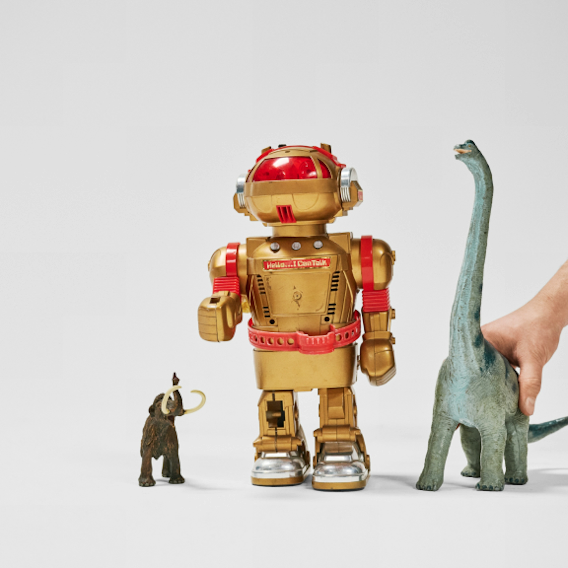 The image shows three toys: a small mammoth figure, a gold robot with red accents, and a large dinosaur figure. A hand is seen holding the dinosaur.