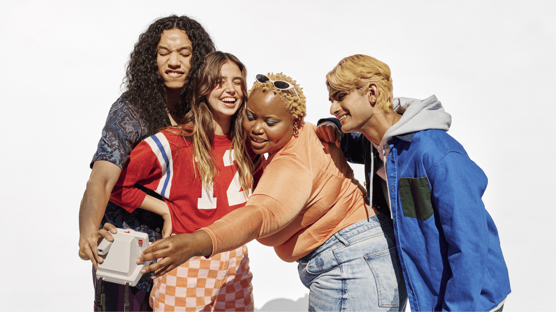 A group of four friends are taking a selfie together, smiling and enjoying each other's company. One person is holding the camera, while the others lean in close. They are dressed in casual, colorful clothing, including a red sports jersey, a blue and green jacket, and a peach-colored top. The background is plain white, putting full focus on their joyful expressions and vibrant outfits.