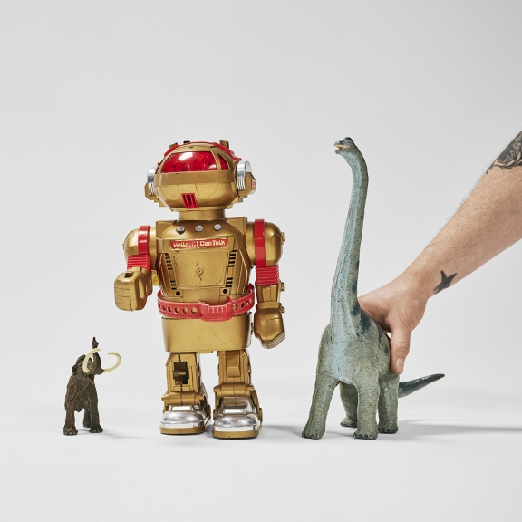 The image shows a toy mammoth, a gold robot, and a dinosaur figure arranged side by side. A hand is holding the dinosaur, which appears to be a Brachiosaurus. The robot has "Hello... I Can Talk" written on its chest.