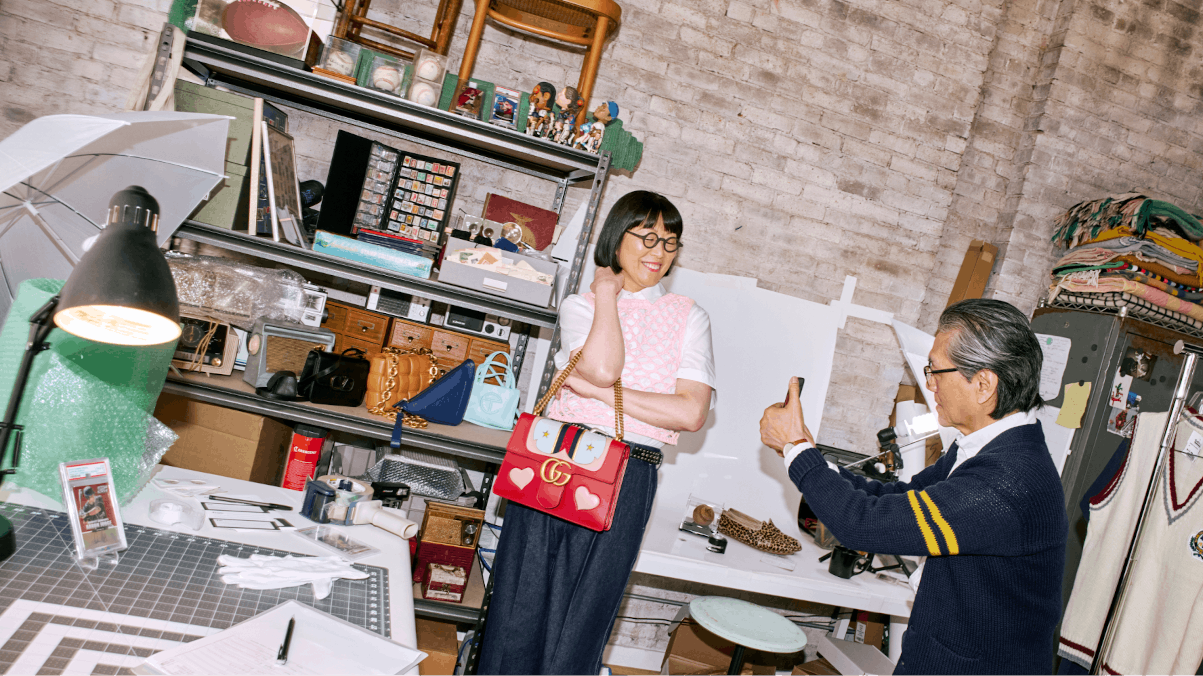 The image shows a vibrant, creative workspace with two individuals—a woman and a man. The woman, smiling and wearing a chic outfit with a colorful handbag, poses while the man takes her photo with his smartphone. The background is filled with various items including lamps, handbags, a sewing station, and shelves stocked with diverse materials, suggesting a lively and dynamic creative environment.