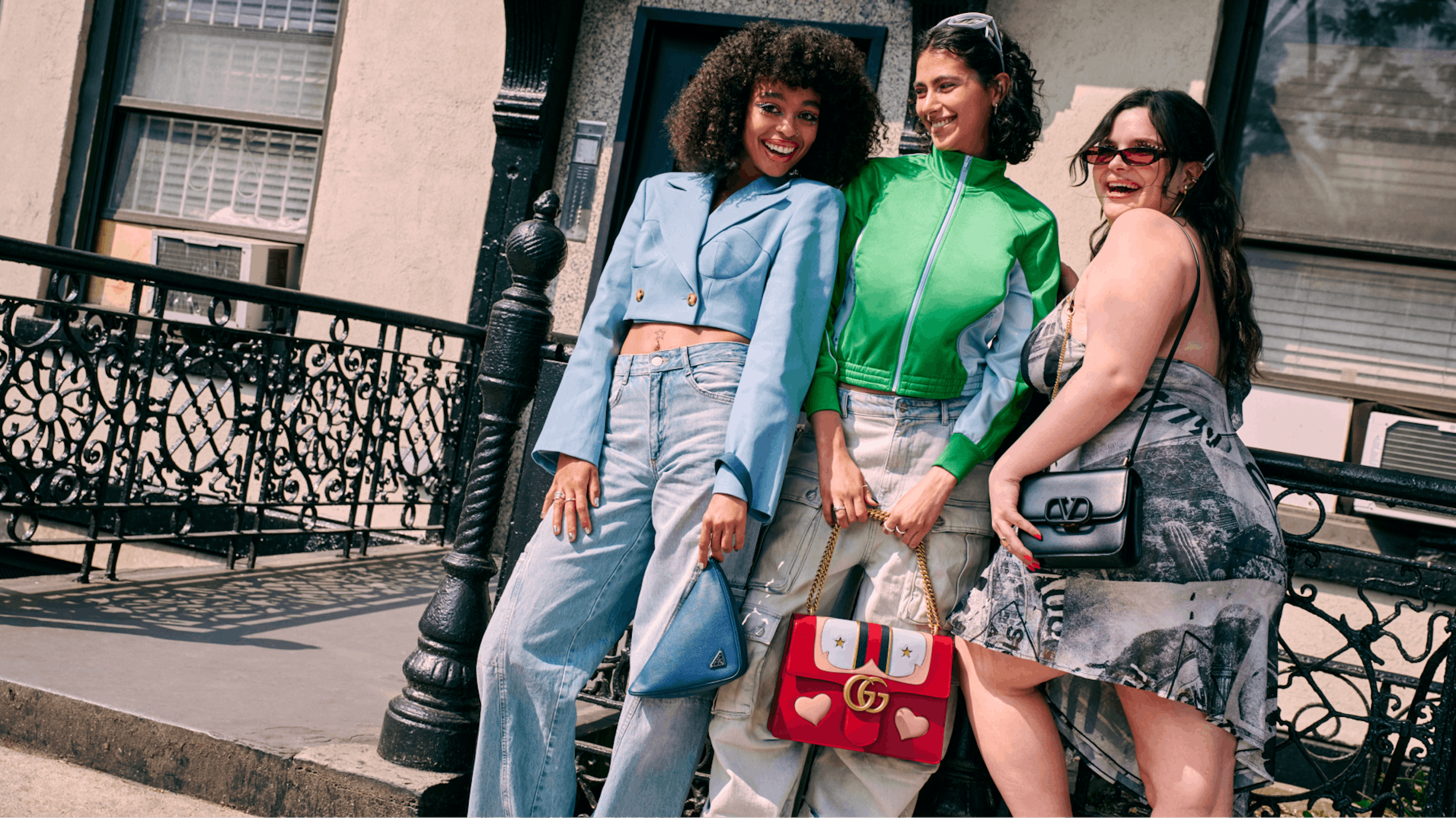 The image shows three women posing on a sunny day by a black iron fence, each showcasing a distinct and stylish outfit. The woman on the left sports a blue cropped blazer and matching jeans, the middle woman wears a vibrant green zip-up jacket, and the woman on the right is dressed in a patterned dress. They each carry fashionable bags, smiling and radiating confidence and camaraderie in a casual urban setting.