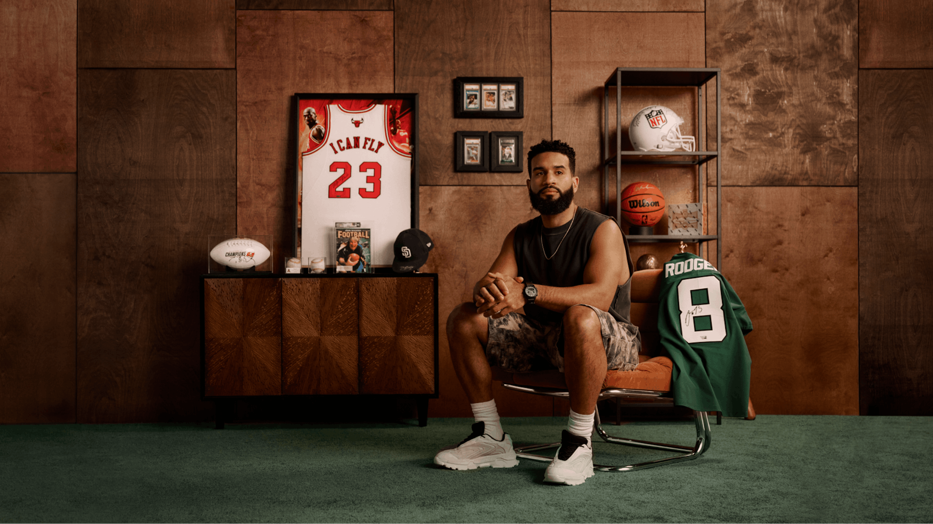 The image shows a man sitting on a stool in a sophisticated, sports-themed room. He's dressed casually in a tank top and camo shorts. Around him are shelves and cabinets displaying sports memorabilia, including basketball and football jerseys, framed pictures, and sports balls. The wooden panel background and green carpet give the room a warm, vintage vibe.