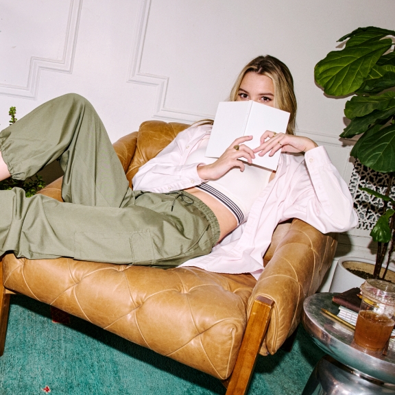 A person is lounging on a tan leather chair, holding a book partially covering their face. They are dressed casually in green pants, a white top, and socks. The setting includes a pink ottoman, a wooden cabinet, a green plant, and a side table with a drink and books, creating a relaxed and cozy atmosphere.