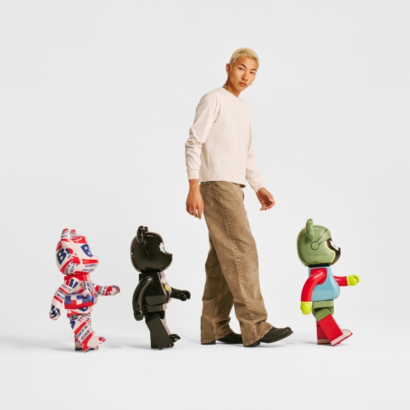 A person with bleached blonde hair, wearing a light long-sleeve shirt and beige pants, is walking alongside three colorful bearbricks. The bearbricks are designed with distinct patterns and colors, creating a playful and whimsical scene against a plain white background.