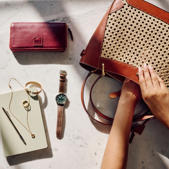 A neatly arranged collection of items is displayed on a marble surface. It includes a red leather wallet, a green-faced watch with a brown strap, a gold bracelet, a gold ring, a necklace, a notebook with a pen, and a woven handbag with leather trim. A pair of hands is seen reaching into the handbag.