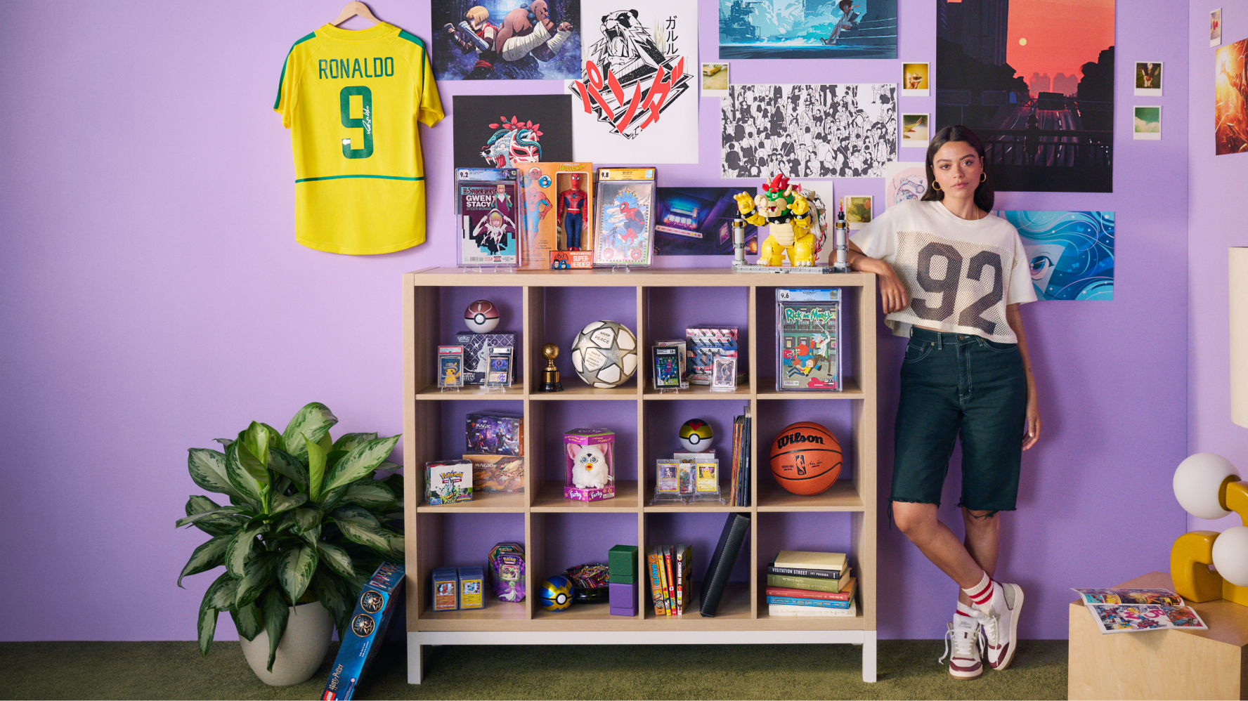 The image displays a young woman standing next to a shelving unit filled with an array of collectibles in a vibrant, colorfully decorated room. The shelves feature items like soccer balls, video games, action figures, and trading cards. A Ronaldo soccer jersey hangs on the wall, alongside various pop culture posters. The woman is casually dressed in a mesh sports jersey and denim shorts, leaning against the shelf in a relaxed pose. The environment reflects a youthful and eclectic mix of interests and hobbies.