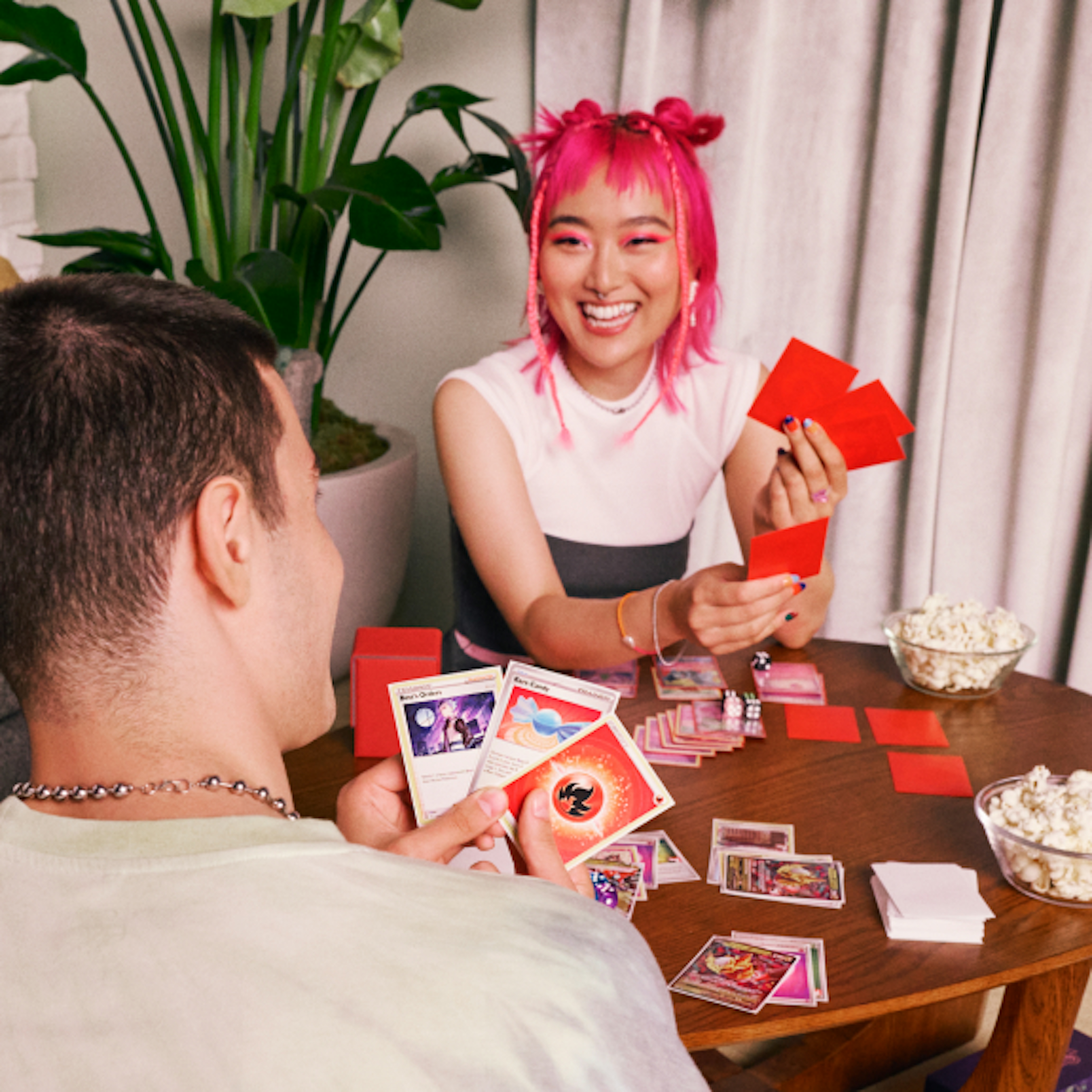 The image captures a lively, indoor gaming scene at home where a woman with vibrant pink hair, is laughing and holding cards in her hand, engaging in a playful card game with a man whose back is to the camera. The table is scattered with more cards and game pieces, and a bowl of popcorn adds to the casual, fun atmosphere. 