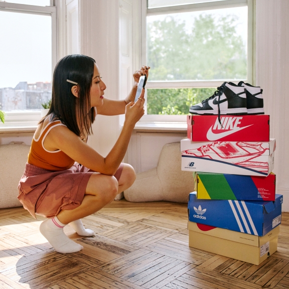 A woman taking a photo of a pair of shoes on top of a stacked of shoe boxes from different companies.