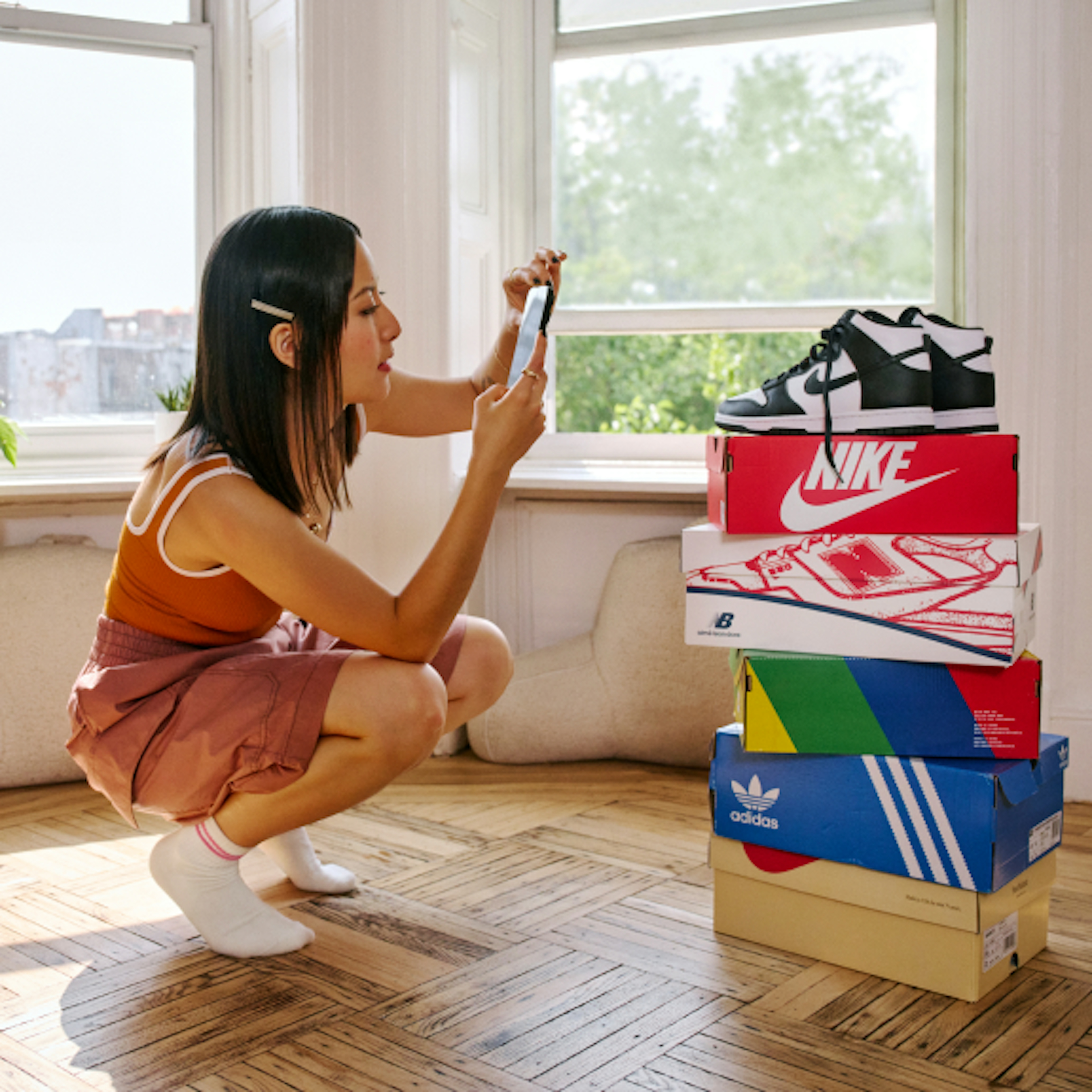 A woman taking a photo of a pair of shoes on top of a stacked of shoe boxes from different companies.